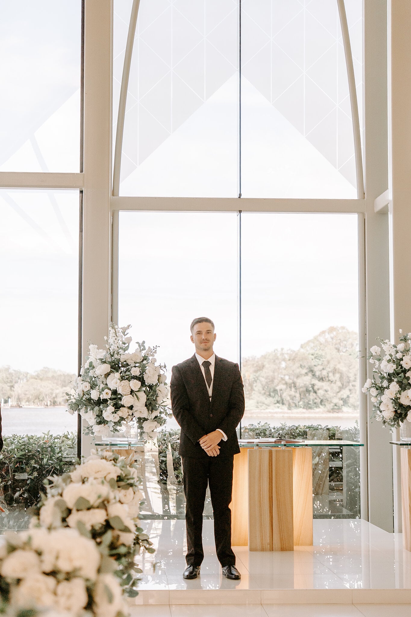 Lane waits at the front of the glass chapel, white florals either side and the Coomera River behind.