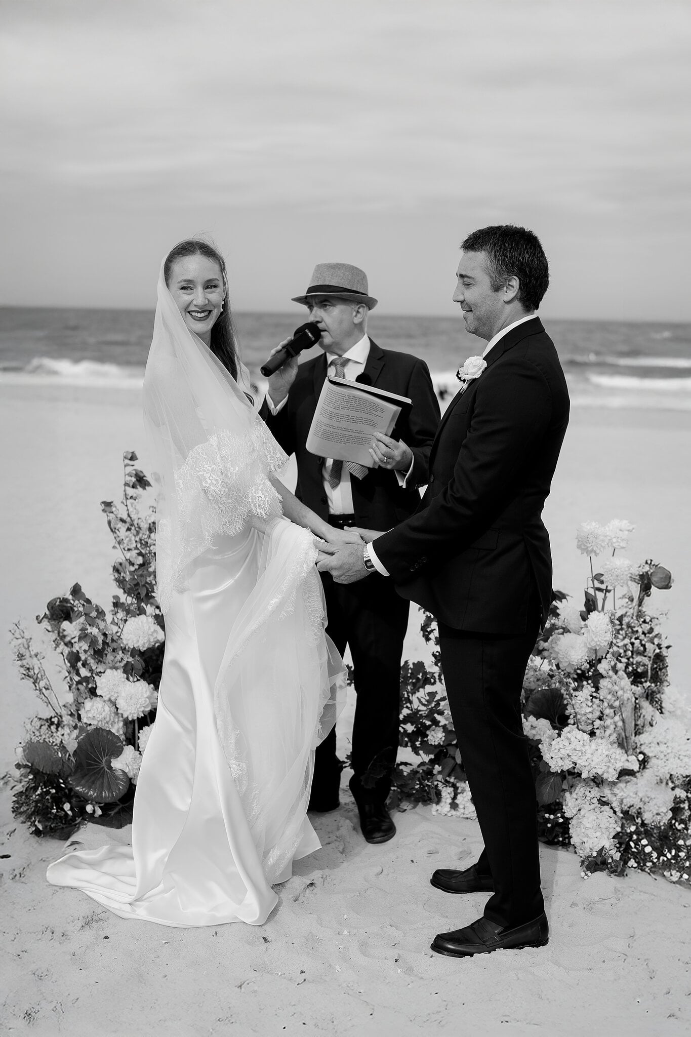 Michele and Nico hold hands for their vows on the sand at South Maroubra Beach, floor florals around them.