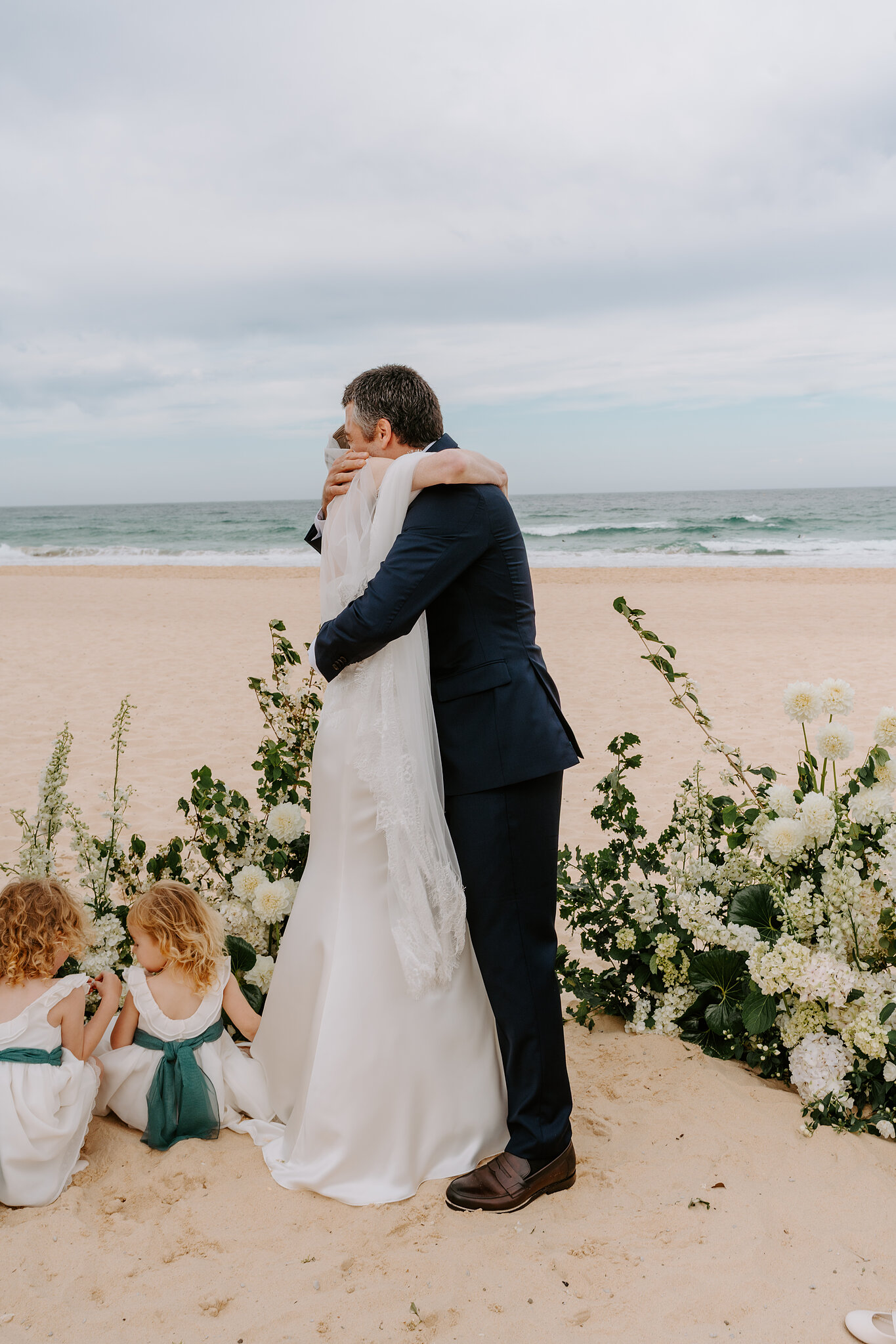 A big hug after the vows at South Maroubra Beach as the flower girls play in the sand beside the florals.