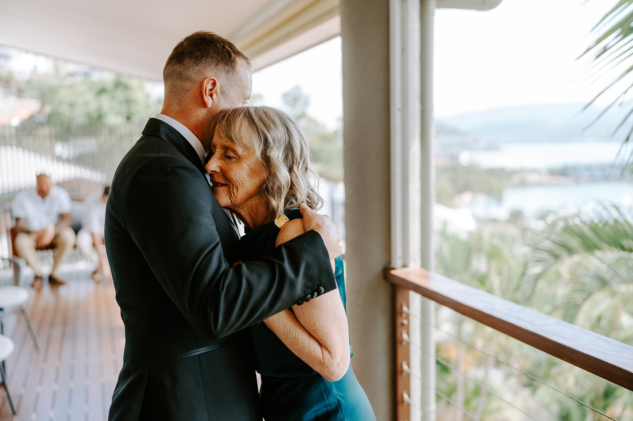 Brendan’s mum hugs him tight on the balcony, the bay behind them.