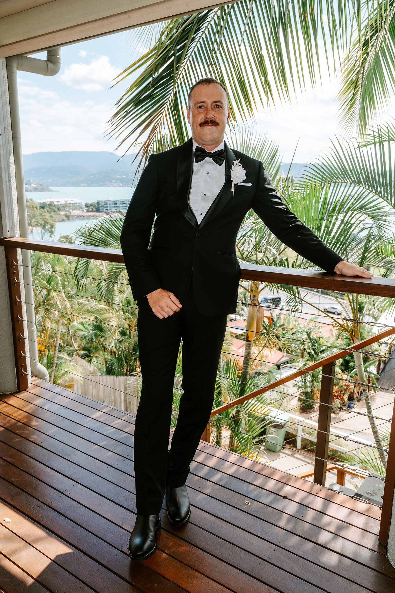 Brendan in his tuxedo on the hillside balcony, palms and the bay behind him.