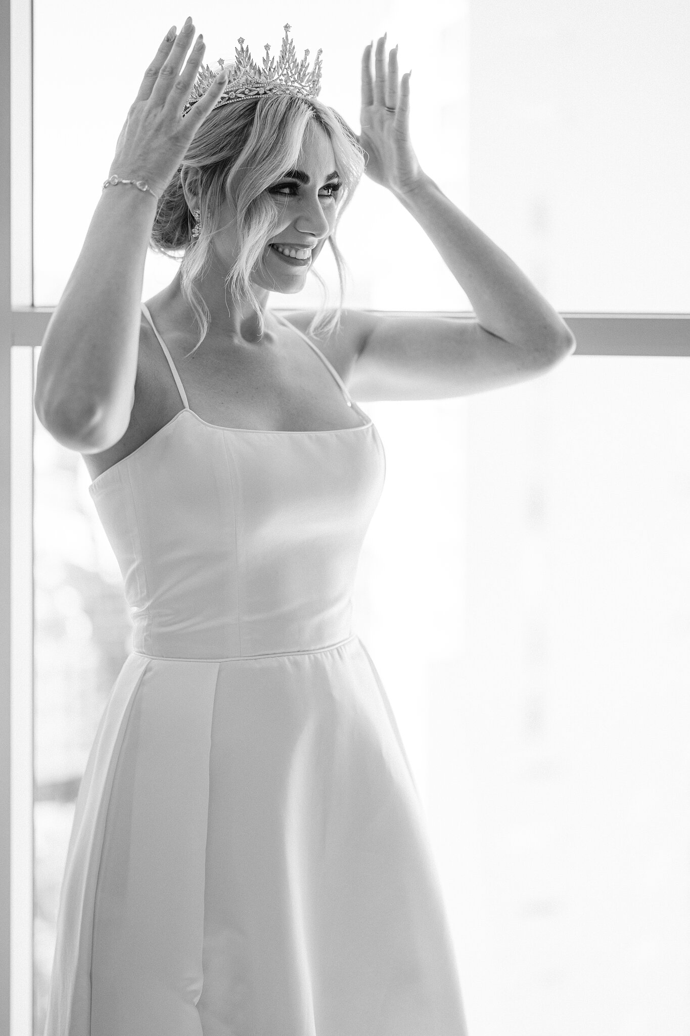 Amanda laughs as she settles a crystal tiara into her hair against a bright window.