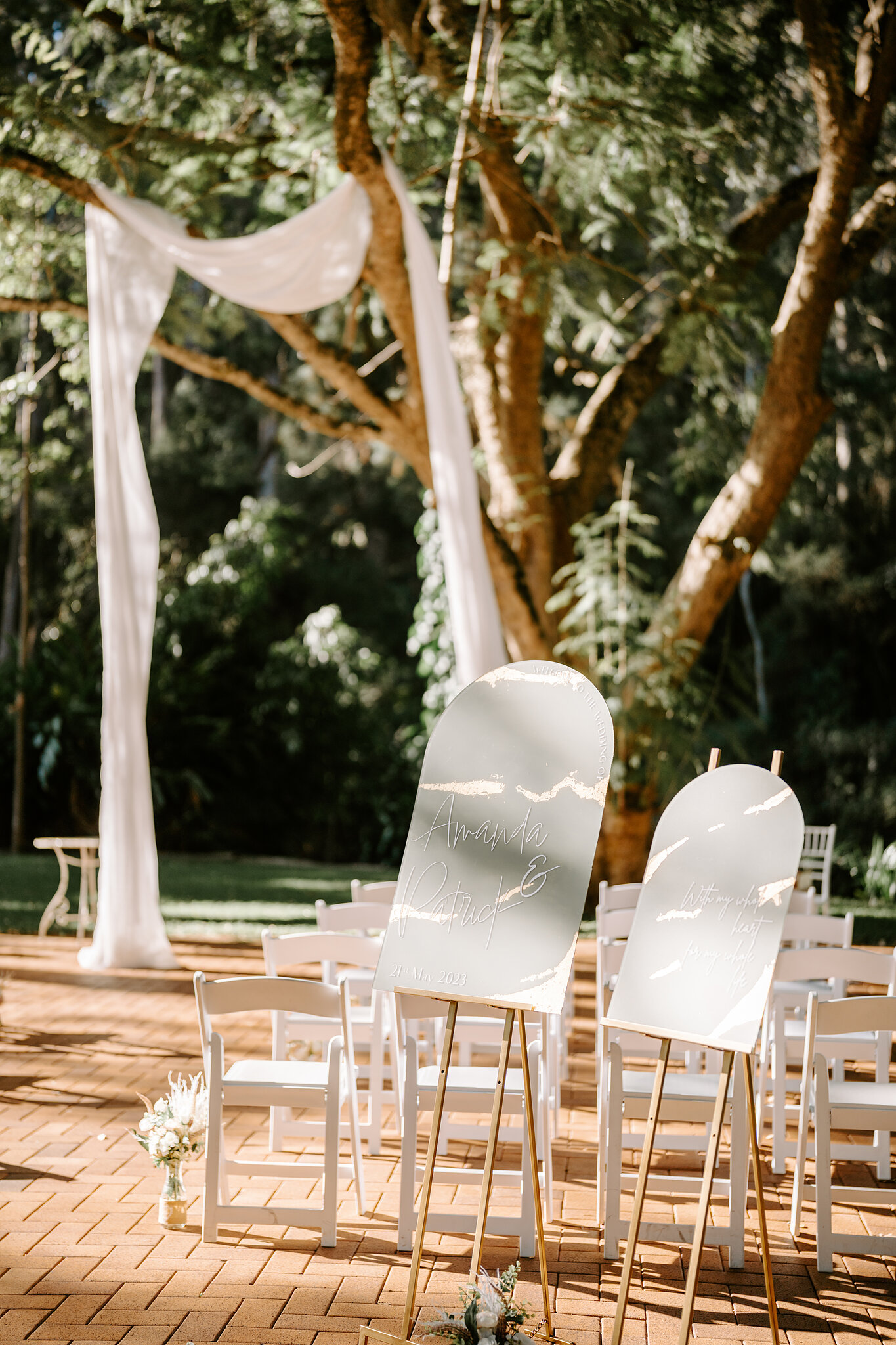 White chairs, draped white silk and arched welcome signs wait on the brick terrace under the Jacaranda Tree at Bundaleer Rainforest Gardens.