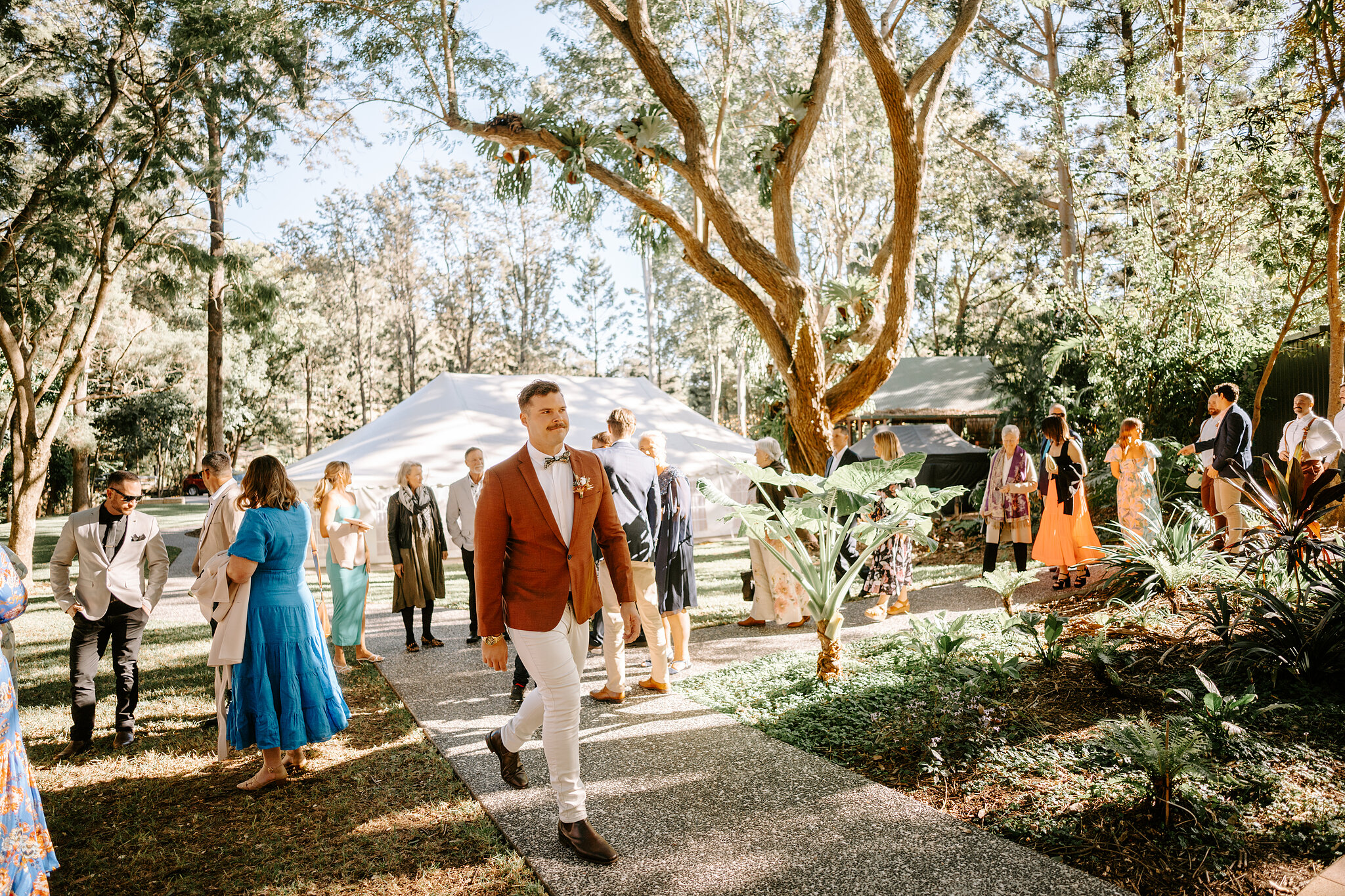Patrick in a rust jacket and bow tie strides up the garden path as guests gather beside the white marquee.