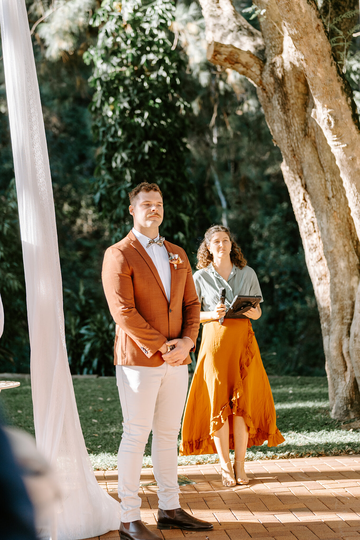 Patrick waits at the front of the ceremony under the trees, hands clasped, watching the aisle.