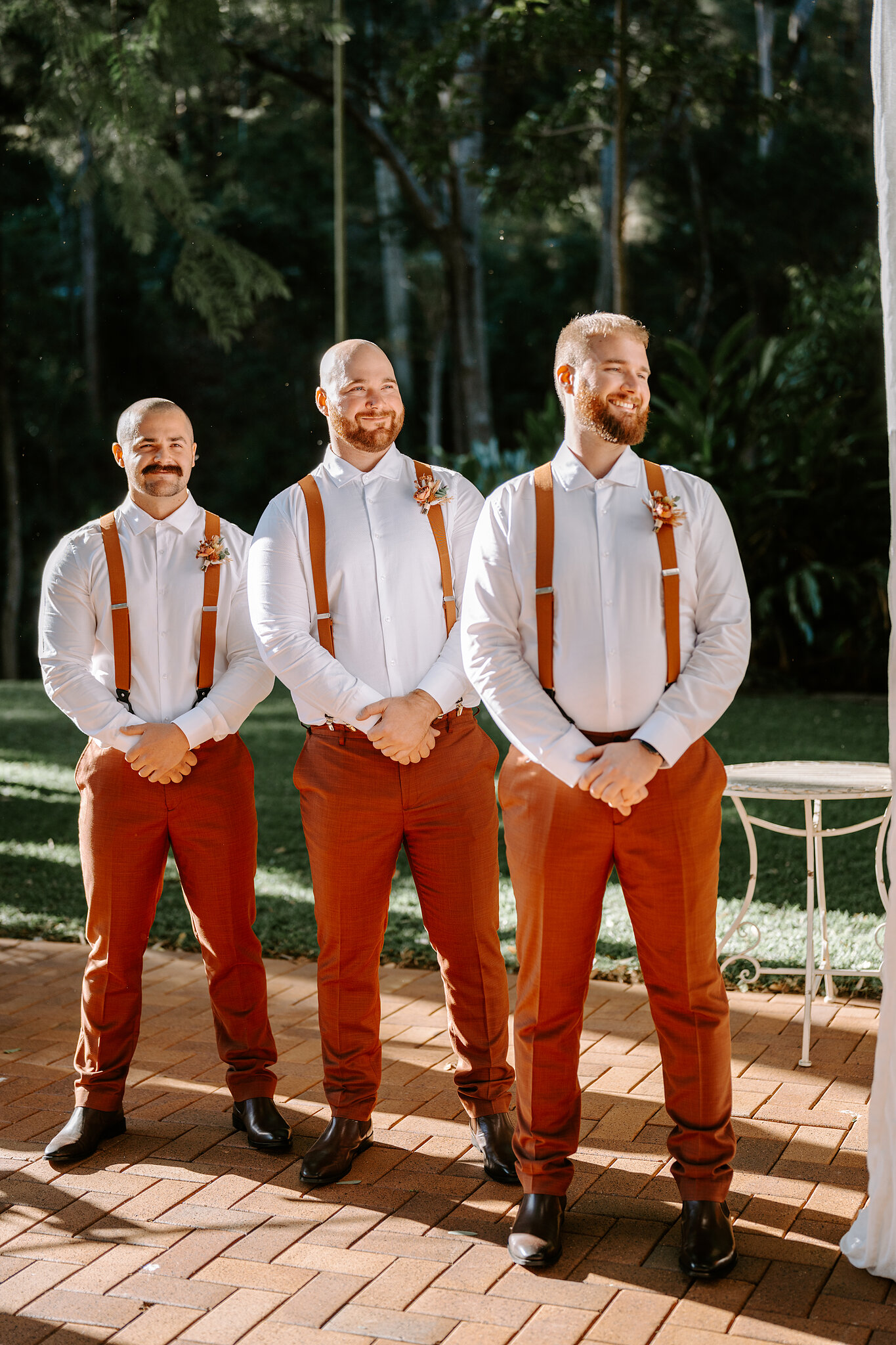 Three groomsmen in rust trousers and braces grin side by side on the brick terrace.