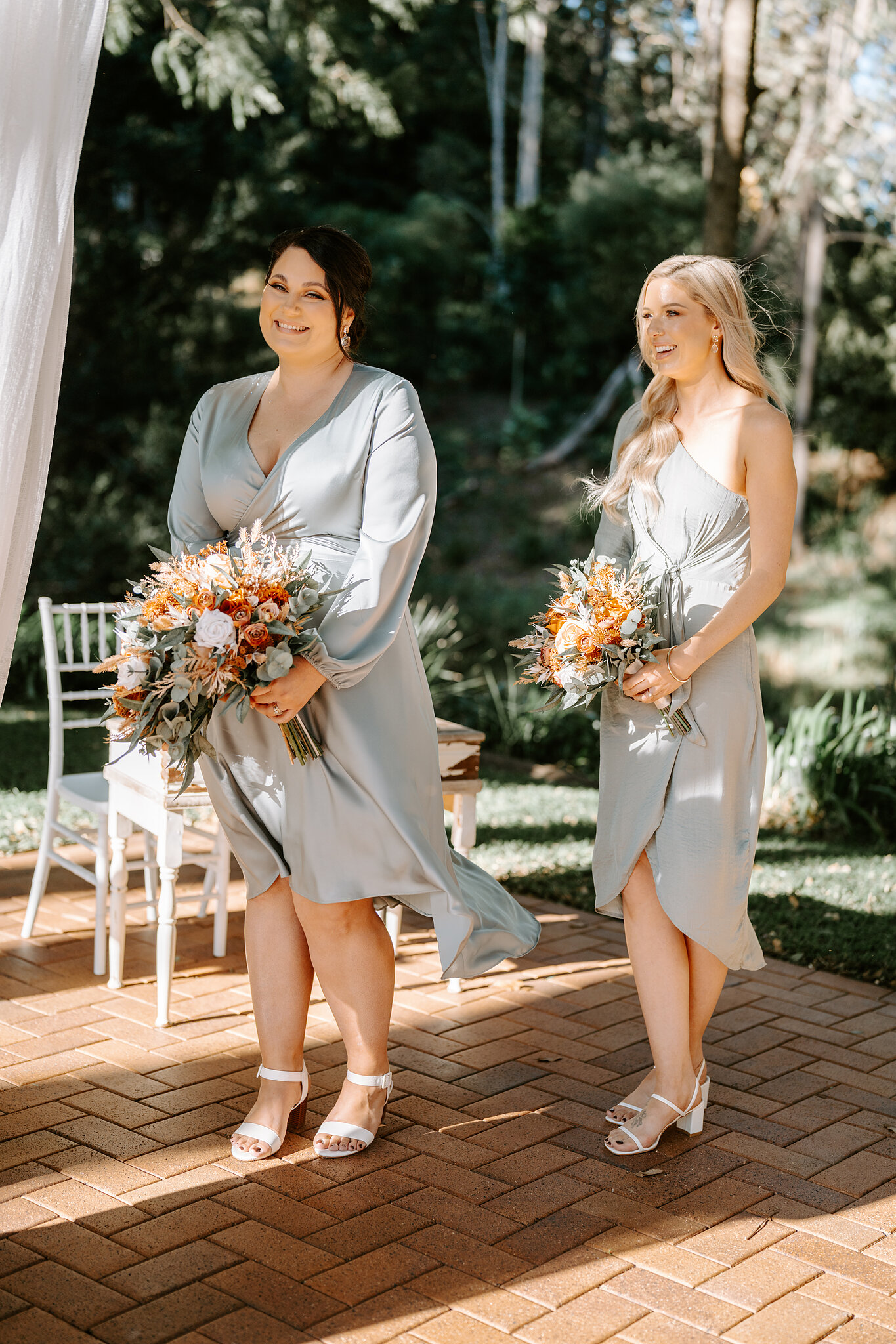 Two bridesmaids in sage satin walk the aisle with bouquets of rust and peach flowers.