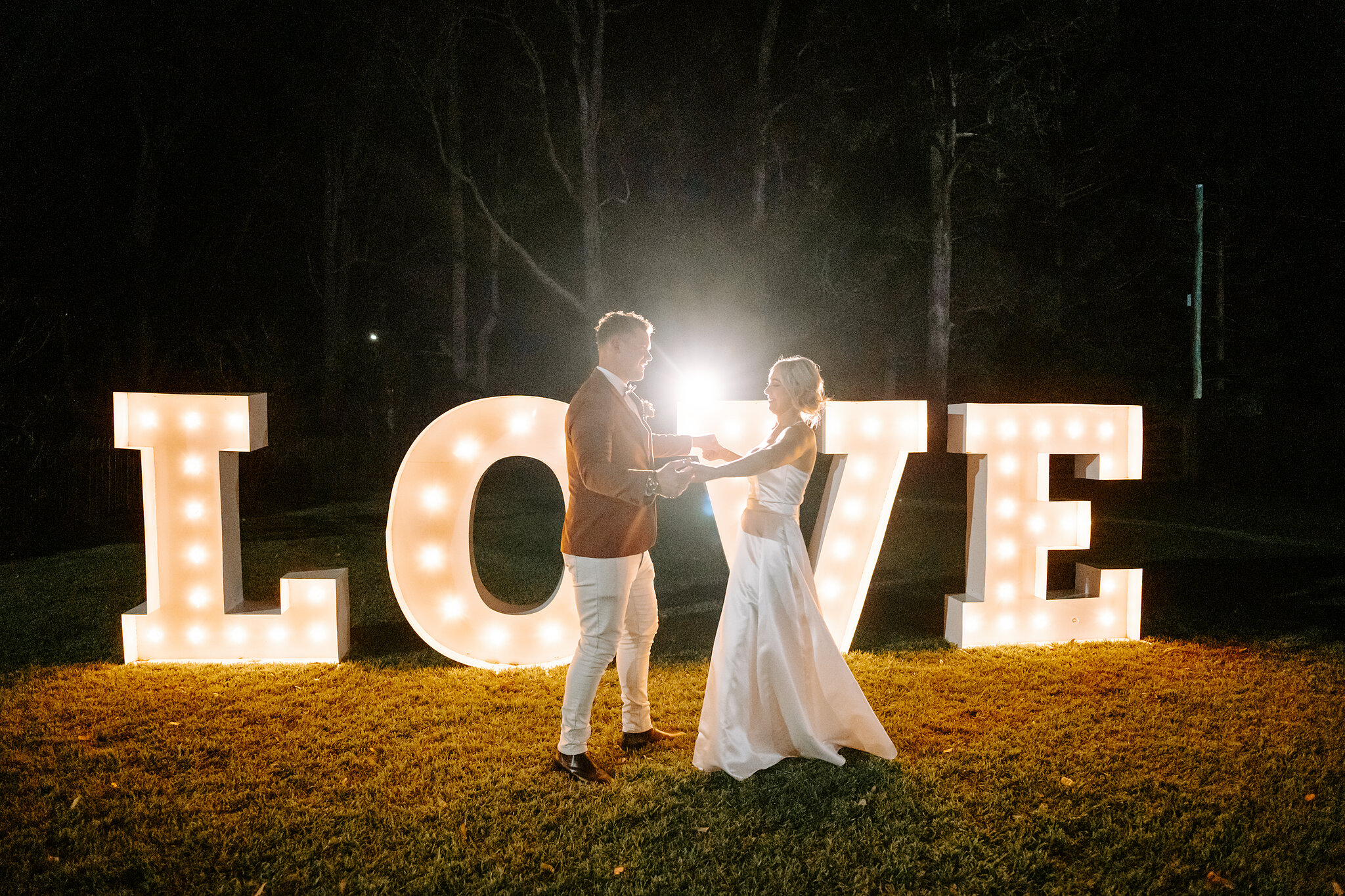 Amanda and Patrick dance hand in hand in front of giant light-up LOVE letters at night.