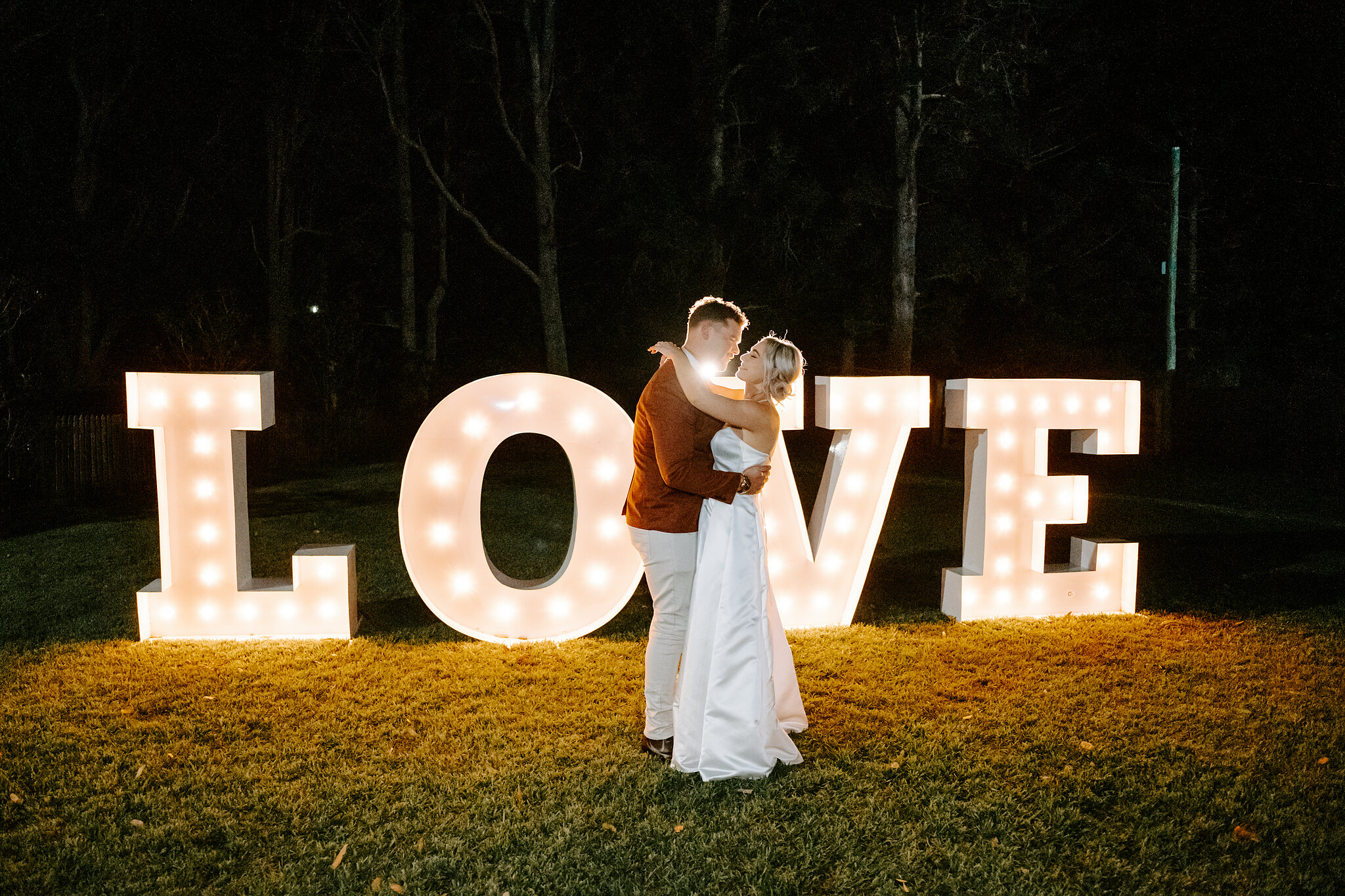 Amanda and Patrick hold each other close in front of the glowing LOVE letters on the lawn.