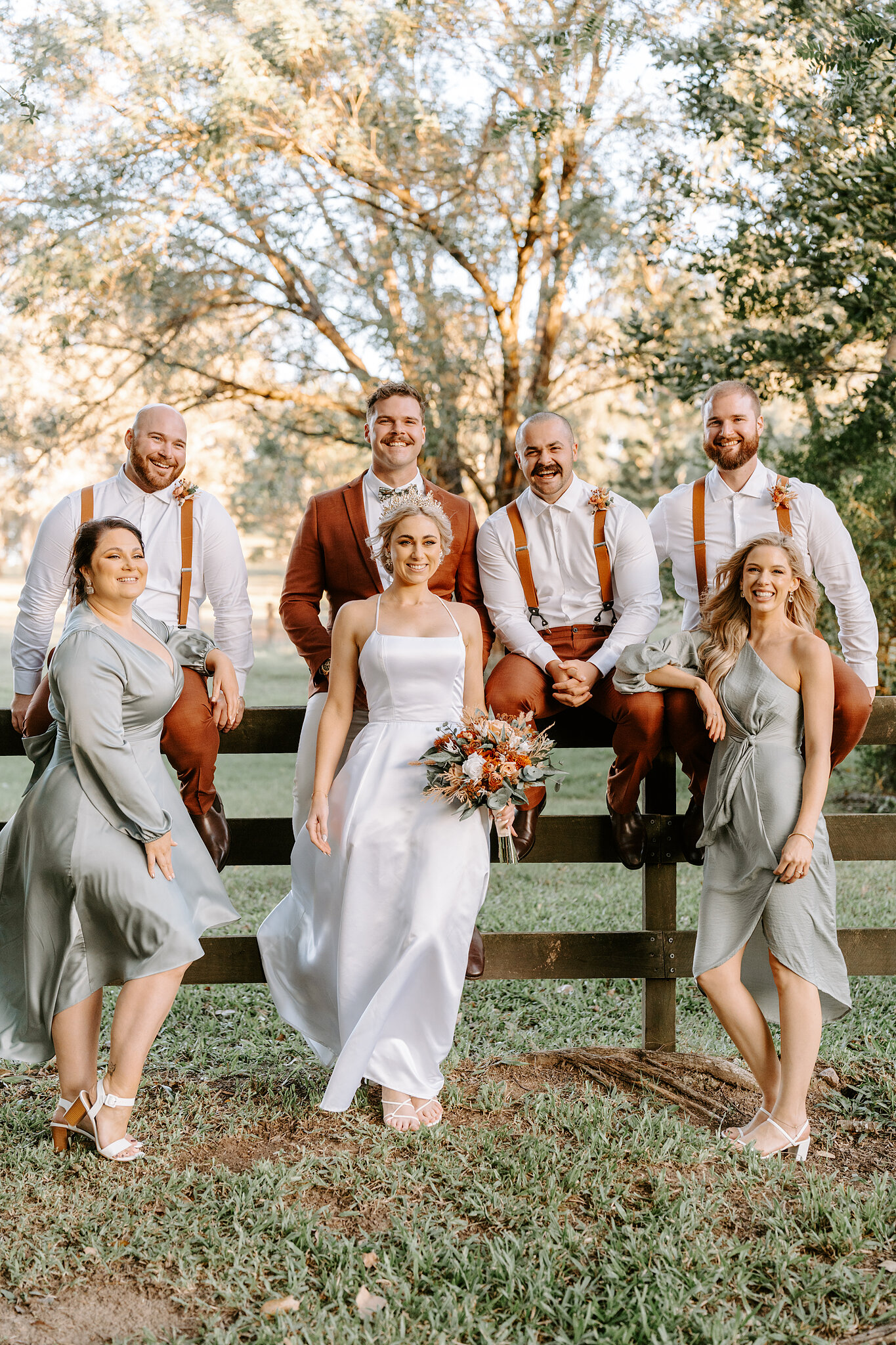 The groomsmen sit on a timber post-and-rail fence behind Amanda, Patrick and the bridesmaids in sage.