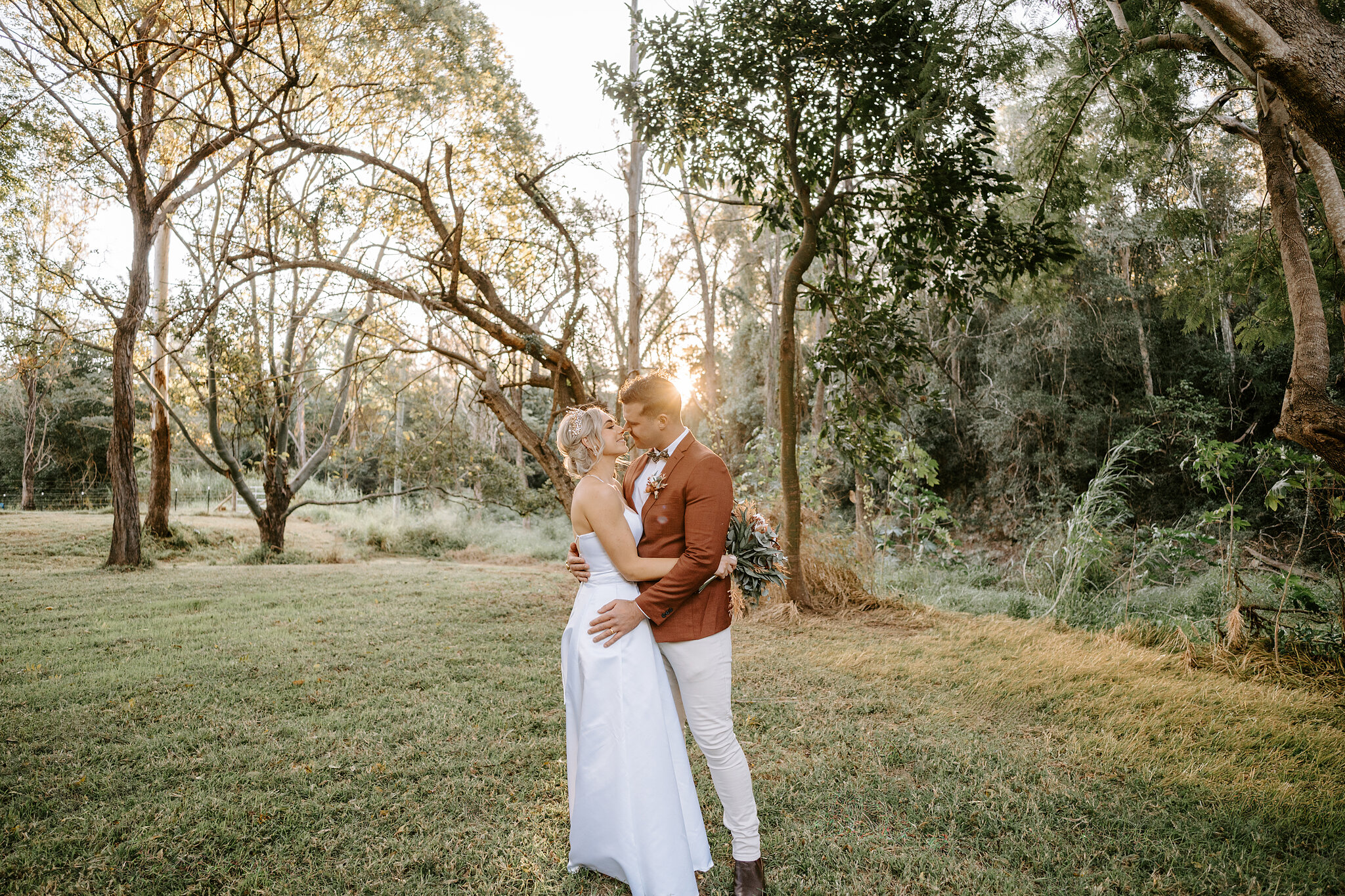 Amanda and Patrick embrace on the lawn at the edge of the rainforest as the sun drops behind the trees.