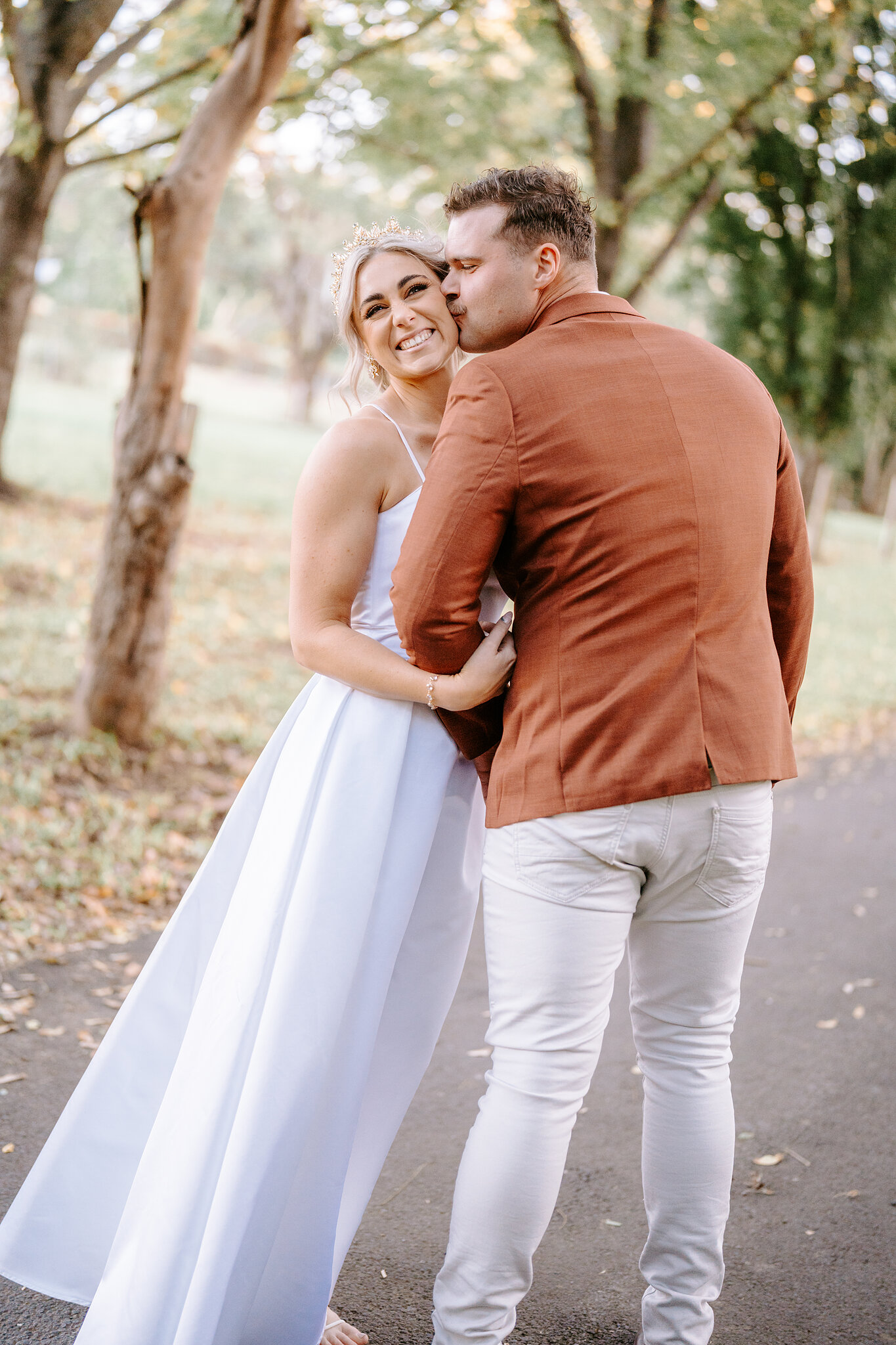 Patrick kisses Amanda’s temple as she grins on the driveway under the trees.
