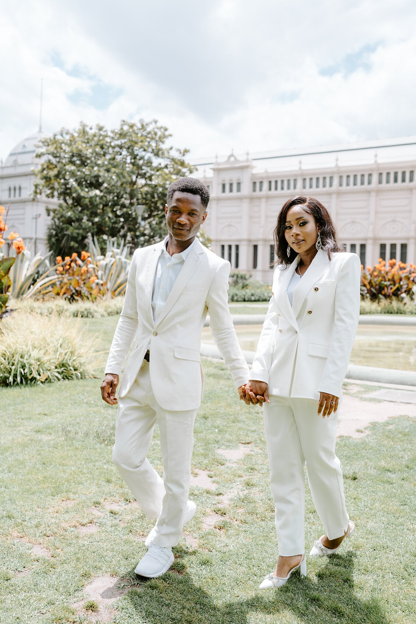 Pearl and Allen walk hand in hand in matching white suits past the Royal Exhibition Building.