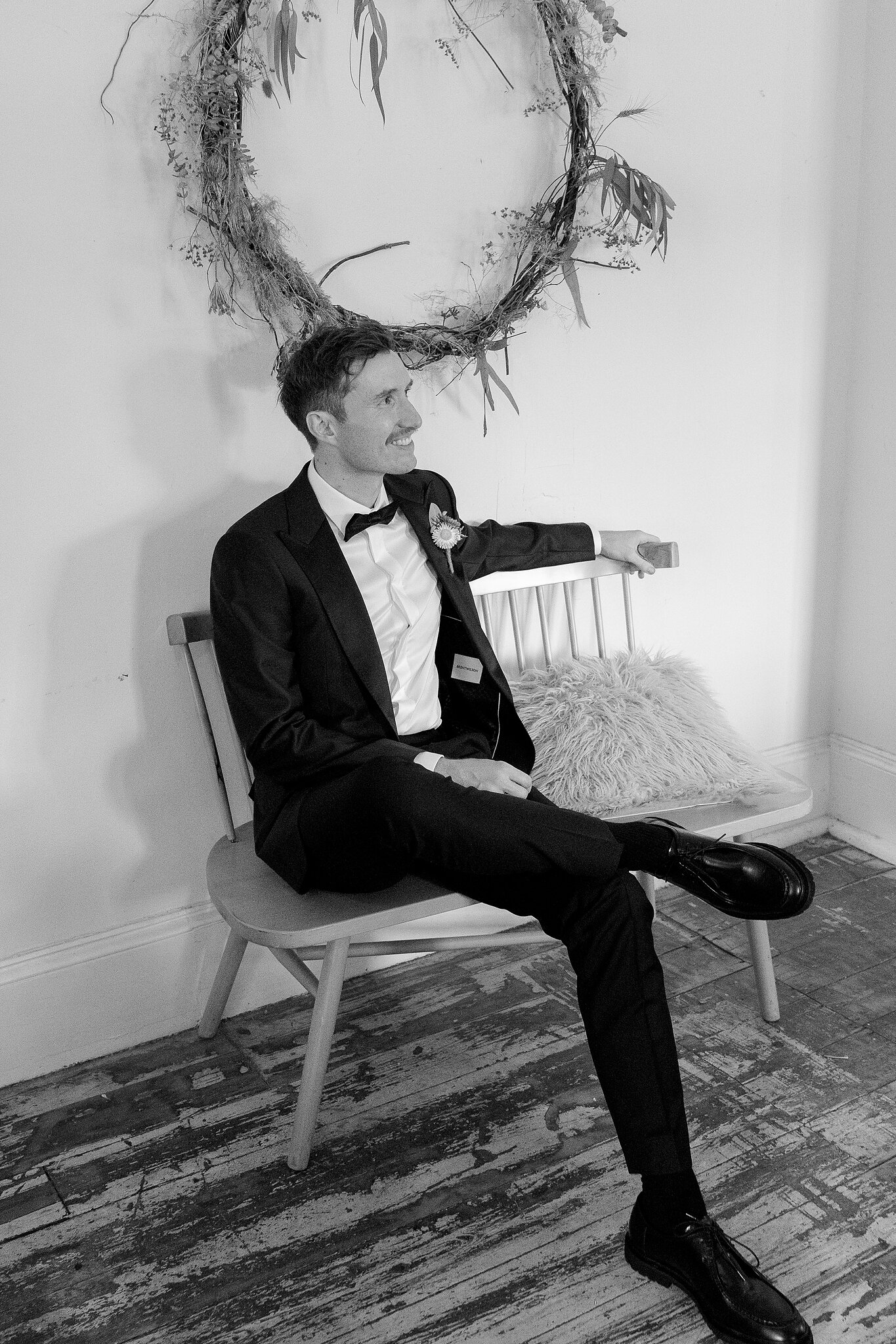 George relaxes on a timber chair under a dried floral wreath, in black and white.