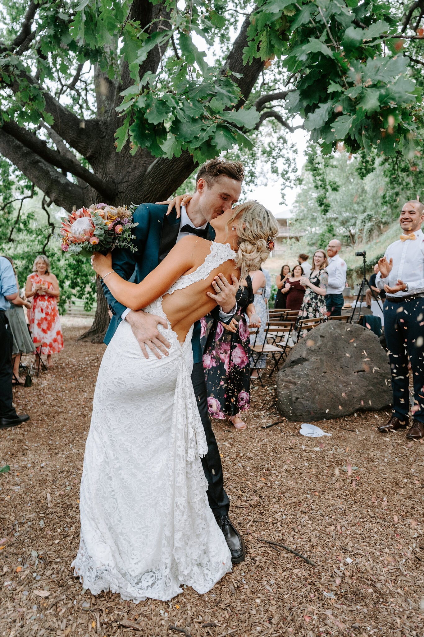 George dips Emma for a kiss under the oak as guests cheer and clap.