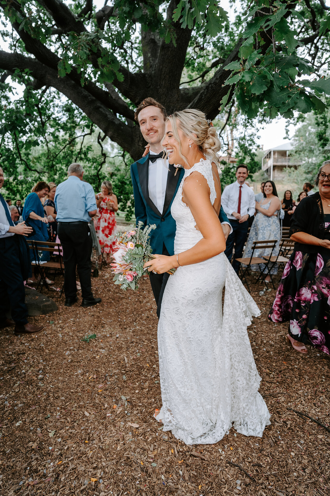 Emma beams beside George as guests clap at the end of the ceremony under the oak.