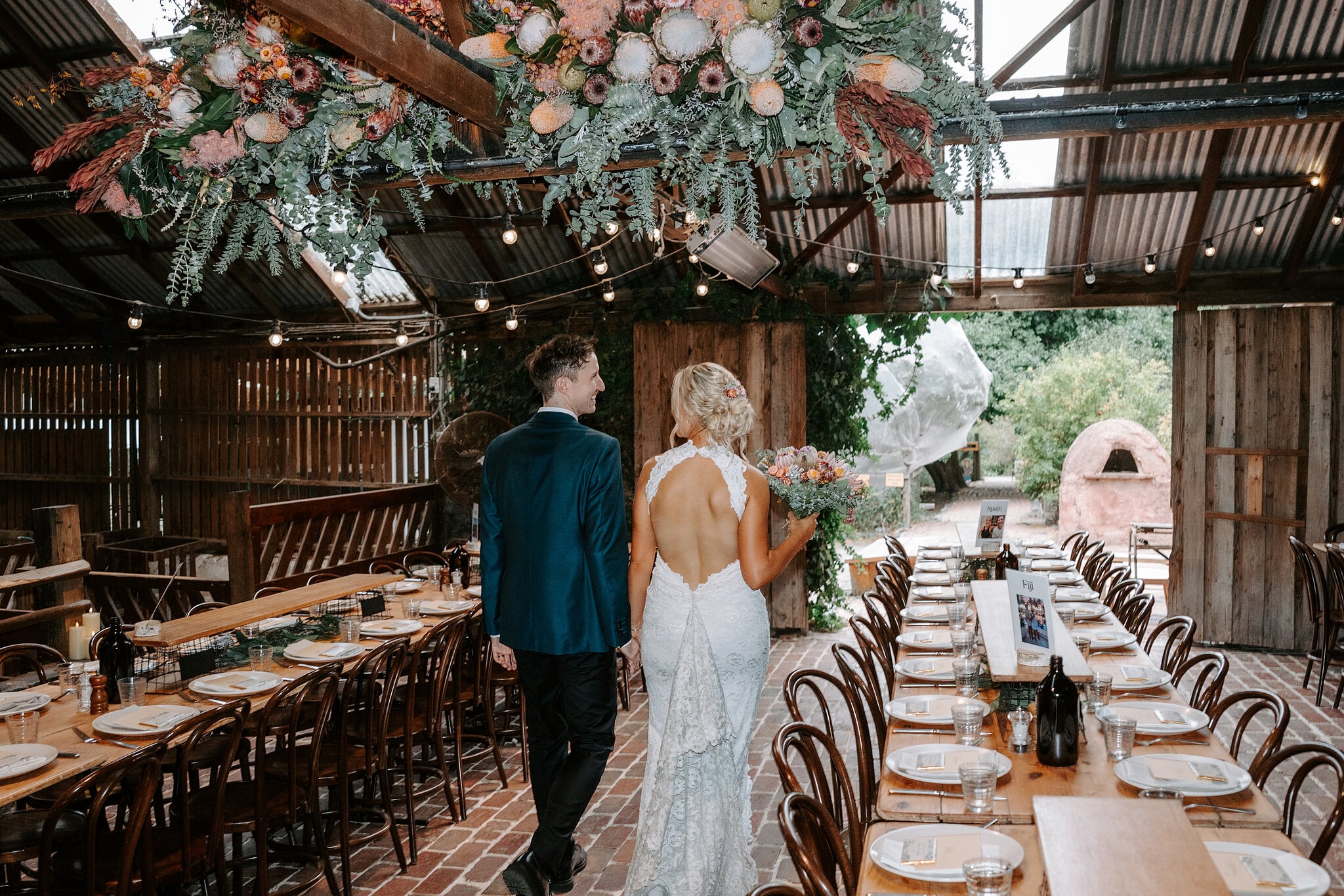 Emma and George step into the Barn, set with long tables under a hanging protea installation.
