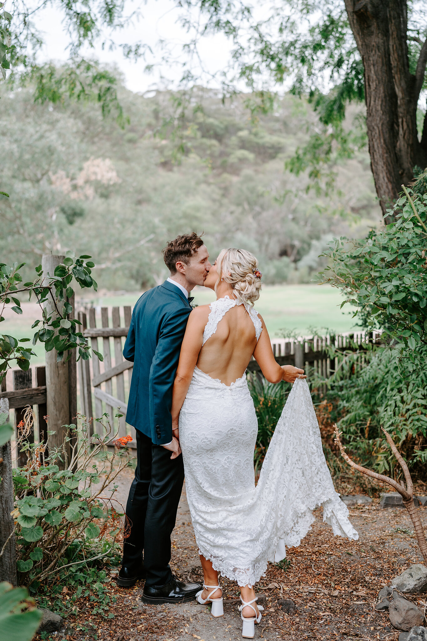 George kisses Emma by a rustic timber fence, her lace train lifted in her hand.