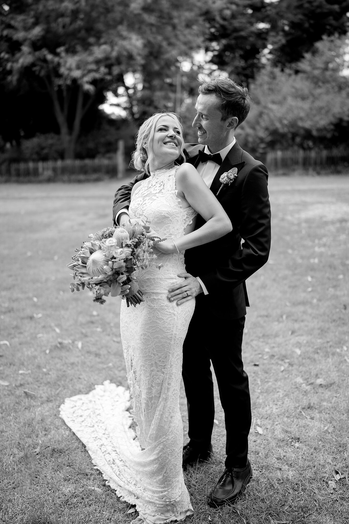 Emma laughs as George hugs her from behind on the farm lawn, in black and white.