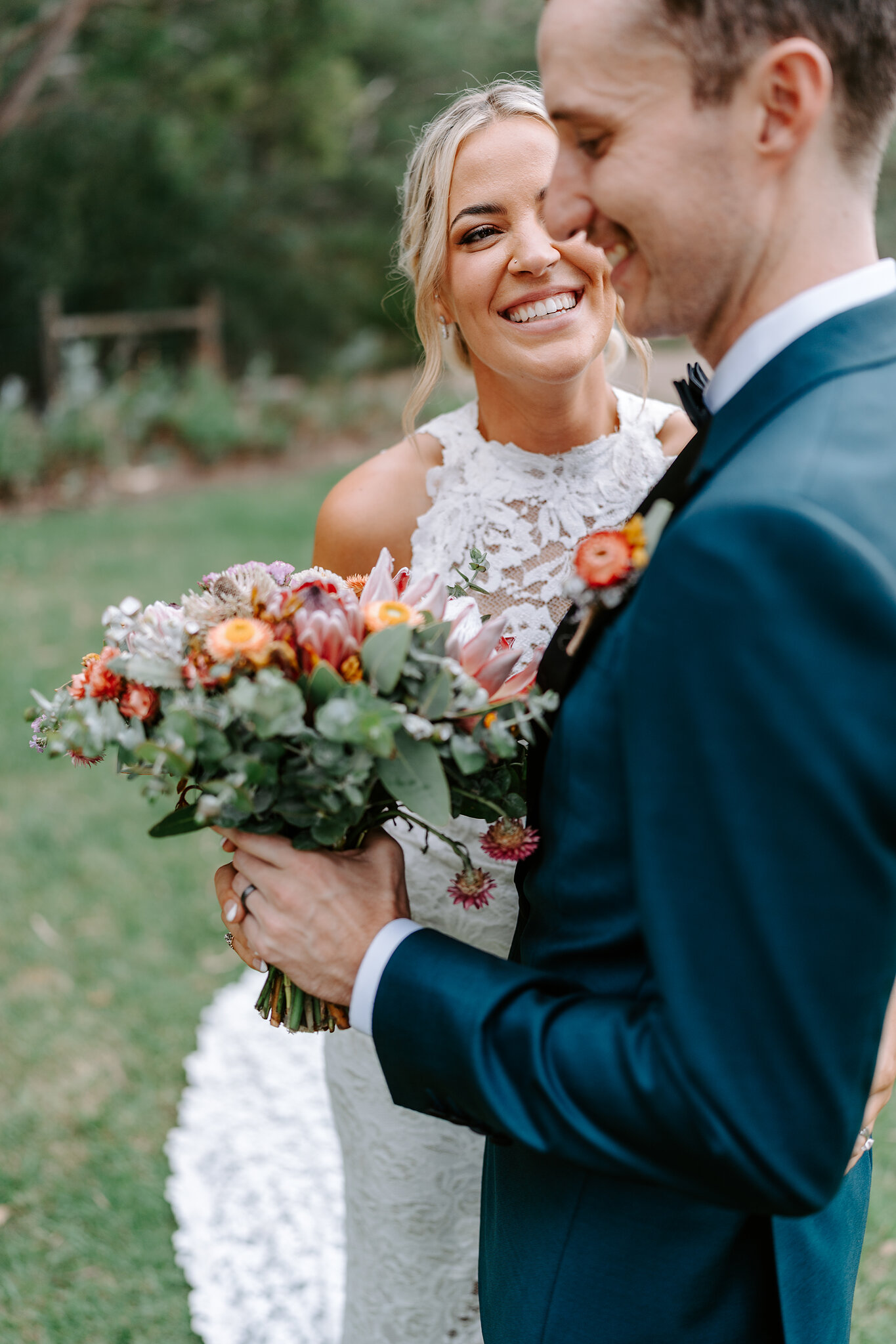 Emma grins into George’s shoulder, her protea bouquet held between them.