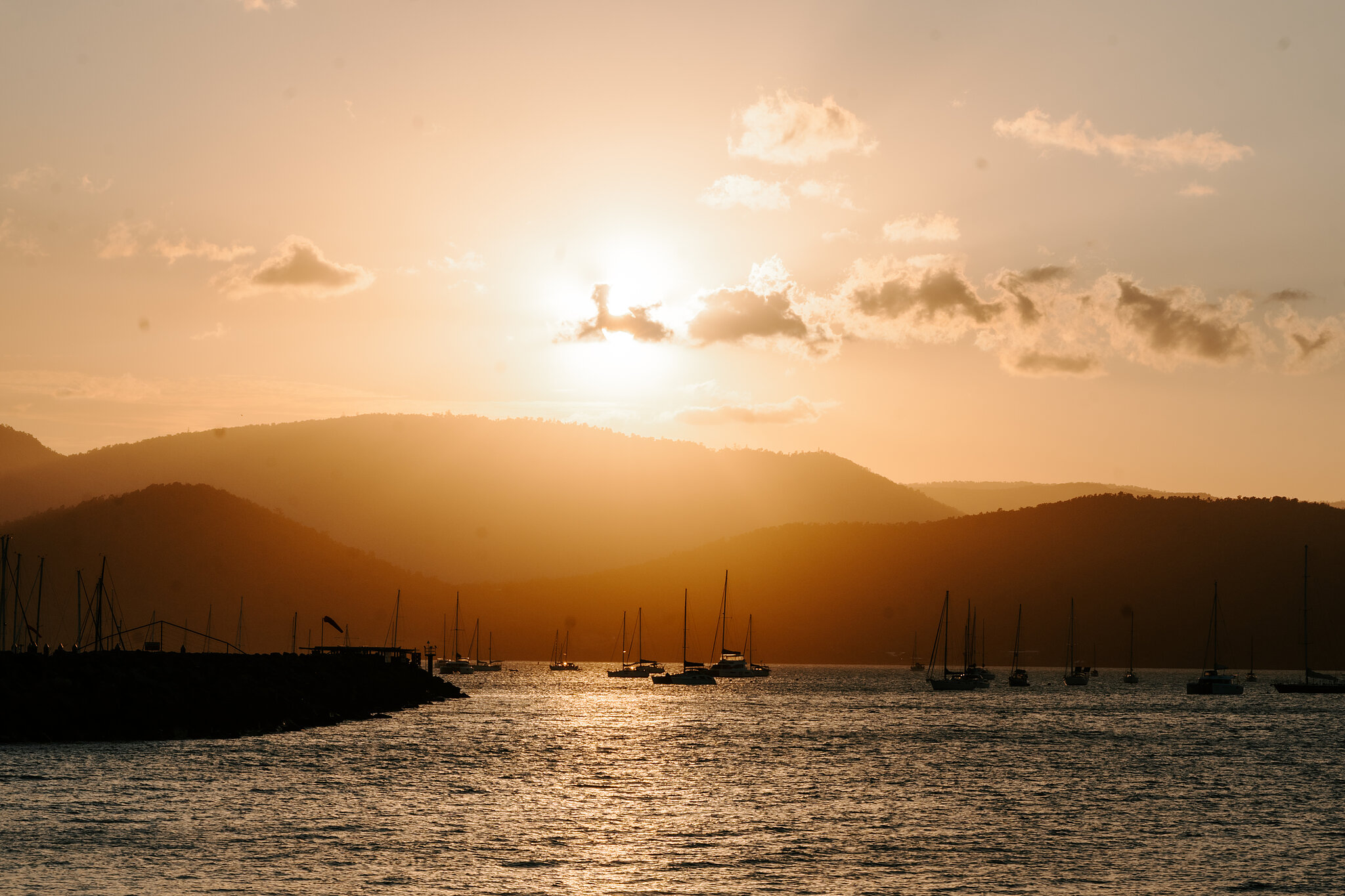 The sun sinks behind the hills across the bay, yachts at anchor below.