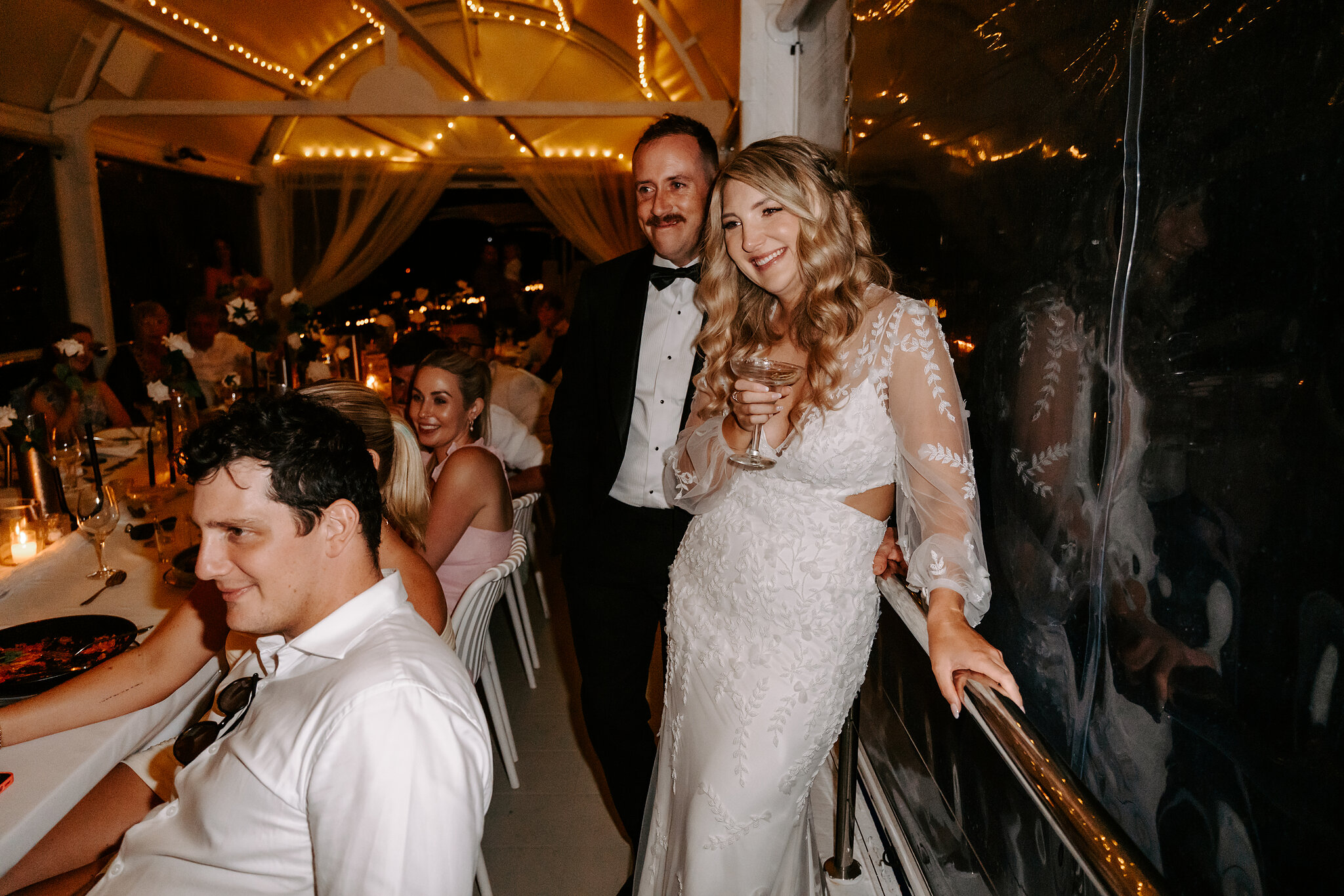 Anna and Brendan laugh with guests at dinner under fairy lights in the jetty marquee.