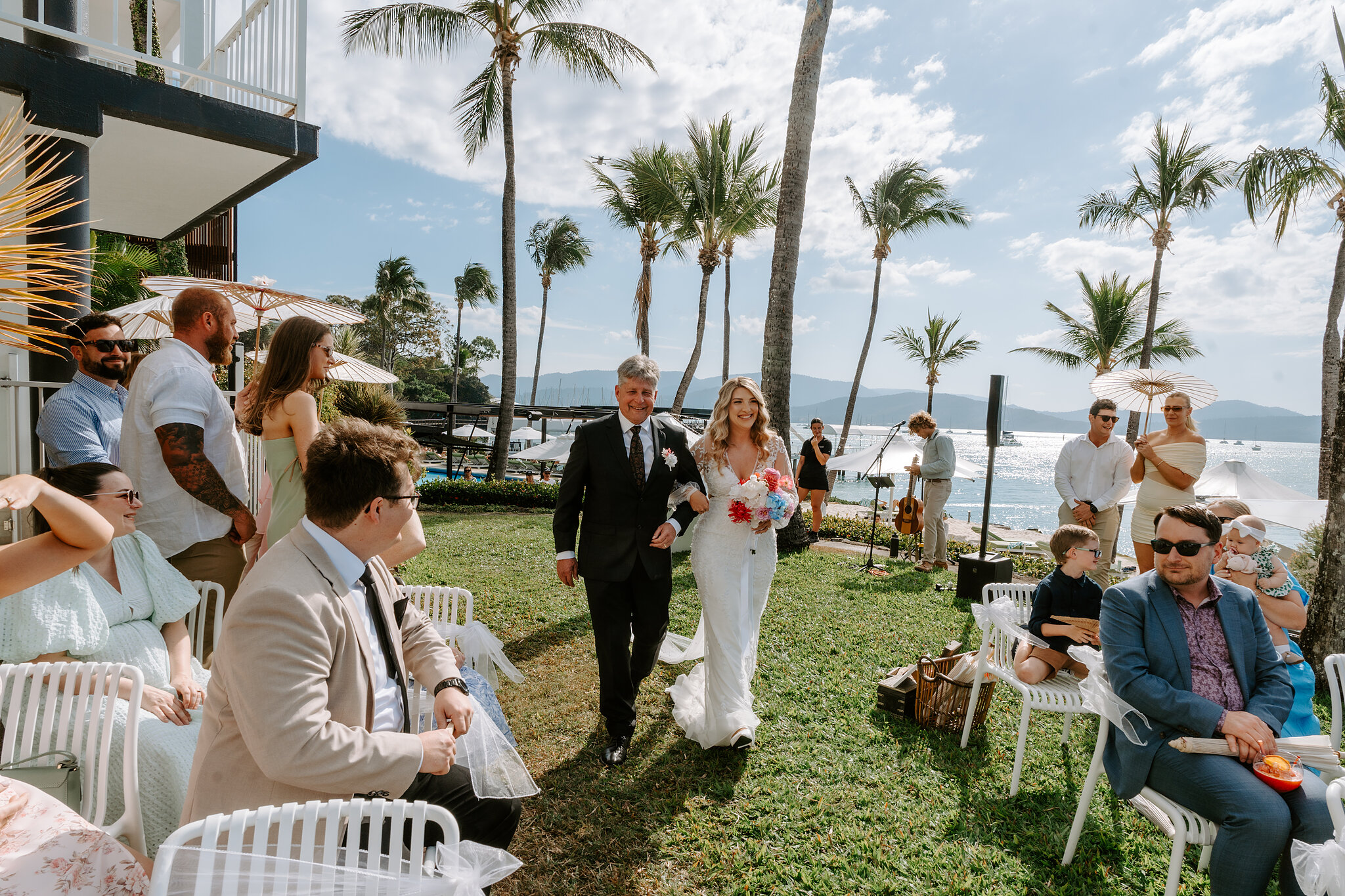 Anna walks down the lawn aisle on her dad’s arm, guests, palms and the sea all around.