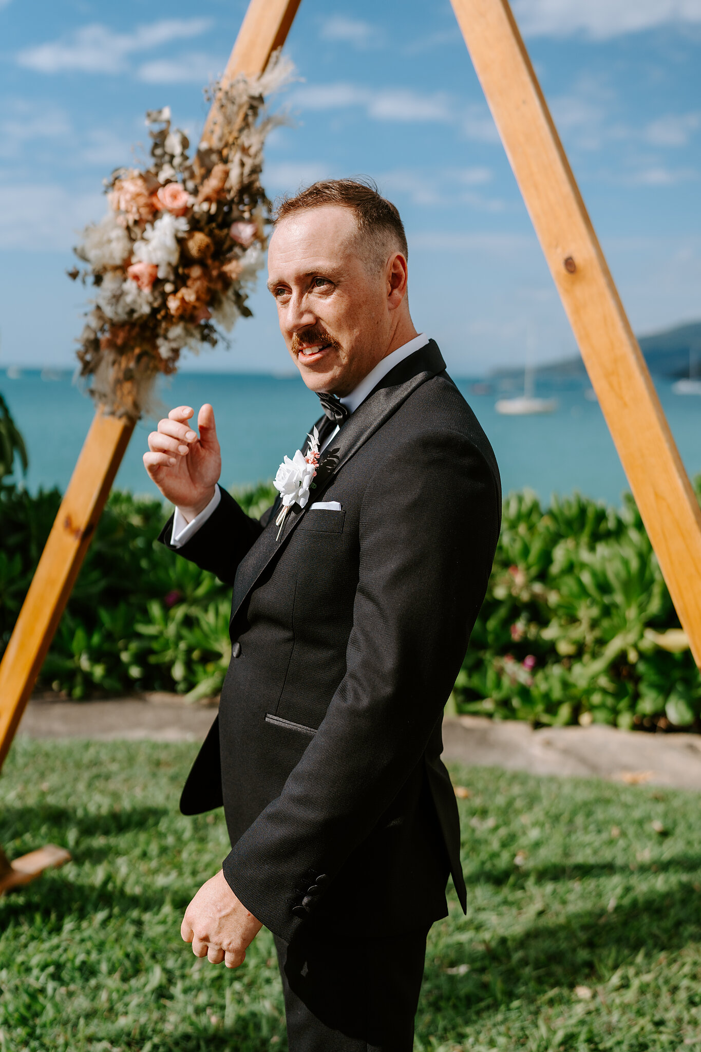 Brendan laughs beside the timber triangle arbour, dried flowers above him and the sea behind.