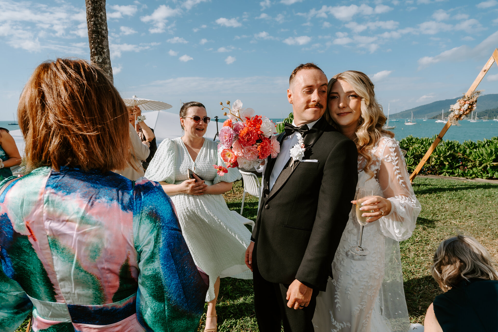 Anna and Brendan cuddle up with champagne as guests congratulate them on the lawn.
