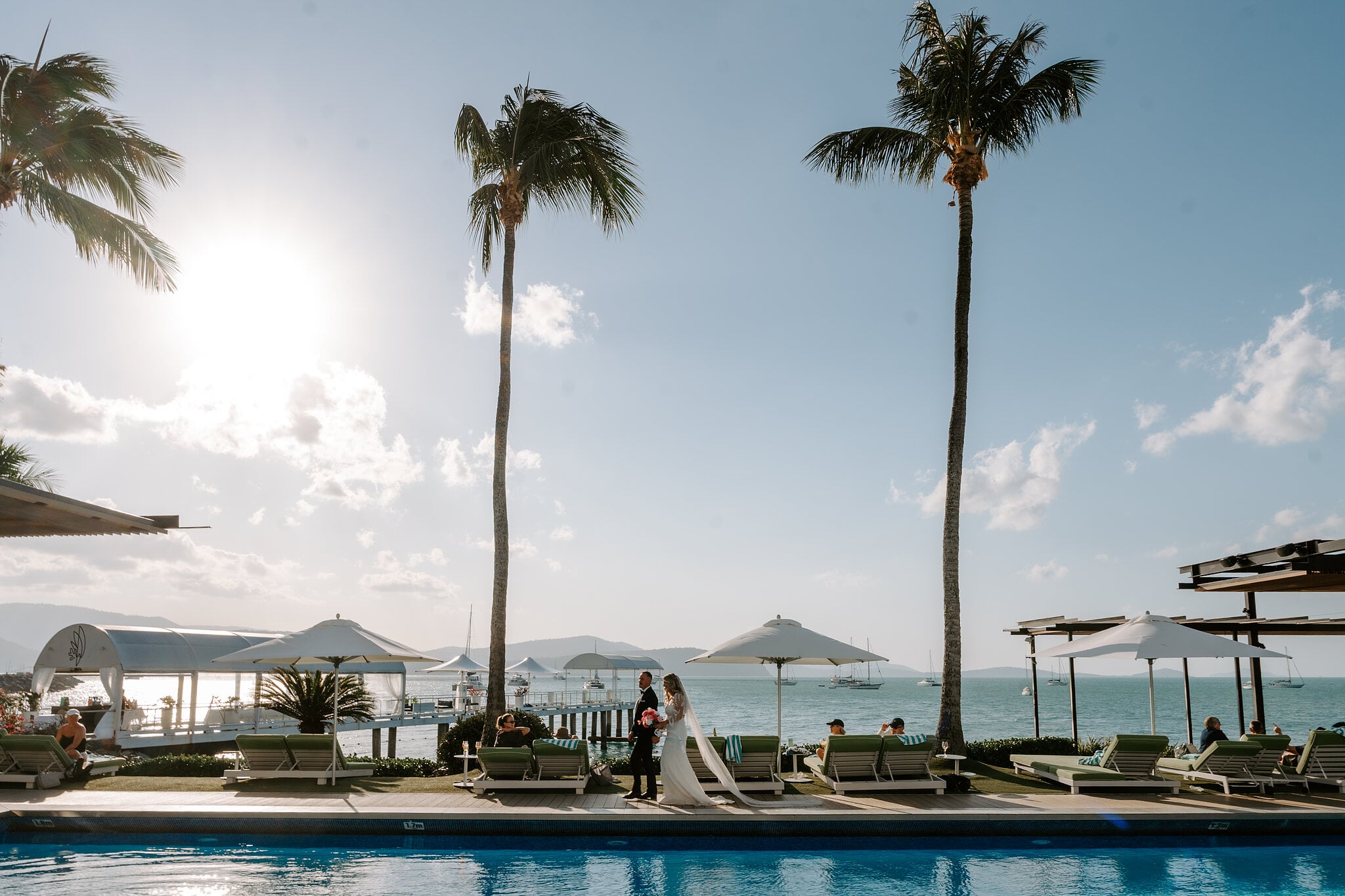 Anna and Brendan cross the pool deck under tall palms, the sun bright over the water.