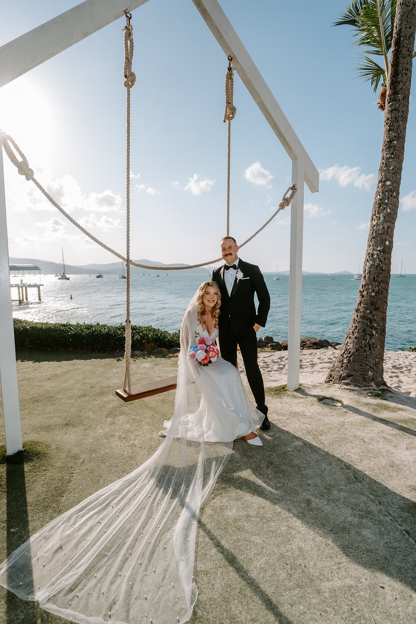 Anna sits on the rope swing by the water with Brendan beside her, her veil spilling across the grass.