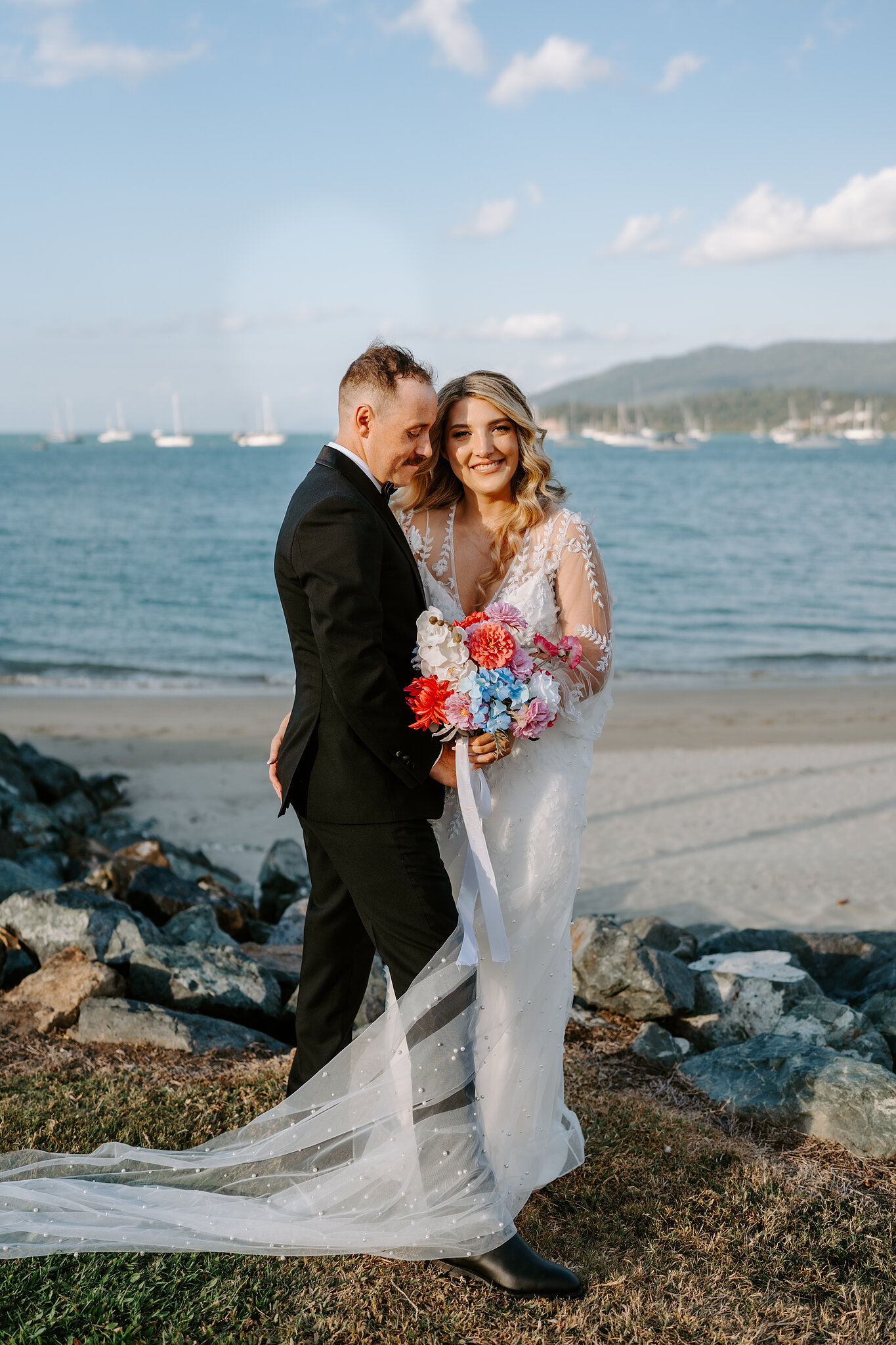 Anna leans into Brendan by the rocks at the water’s edge, her bouquet bright against the sea.