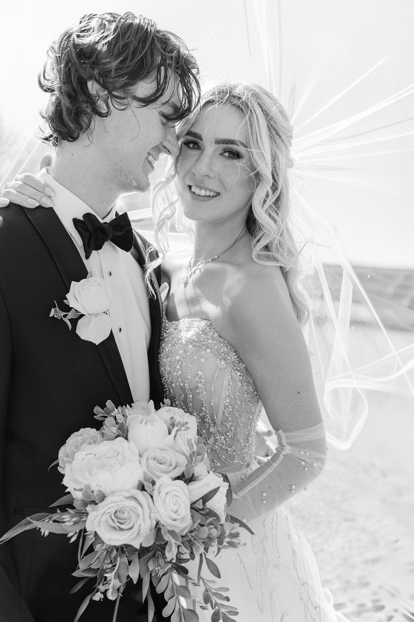 Black and white of Claire and George cheek to cheek on the sand, her veil lifting.