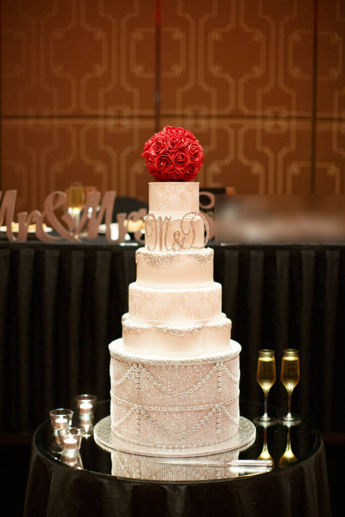 A tall white tiered cake crusted in crystals and topped with a ball of red roses waits in the ballroom at Palladium at Crown