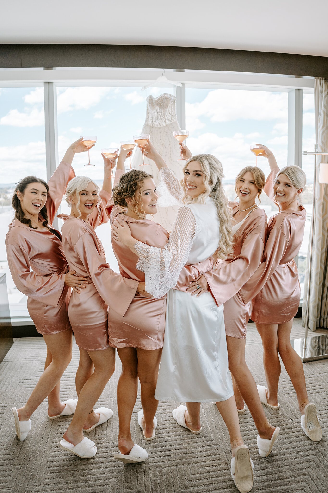 Claire and her bridesmaids in blush satin robes raise coupes of bubbly by the window.