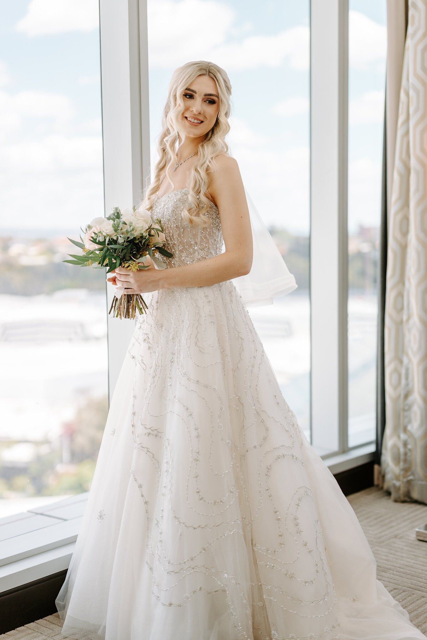 Claire holds her blush and ivory bouquet by the tall windows at Crown Towers Perth.