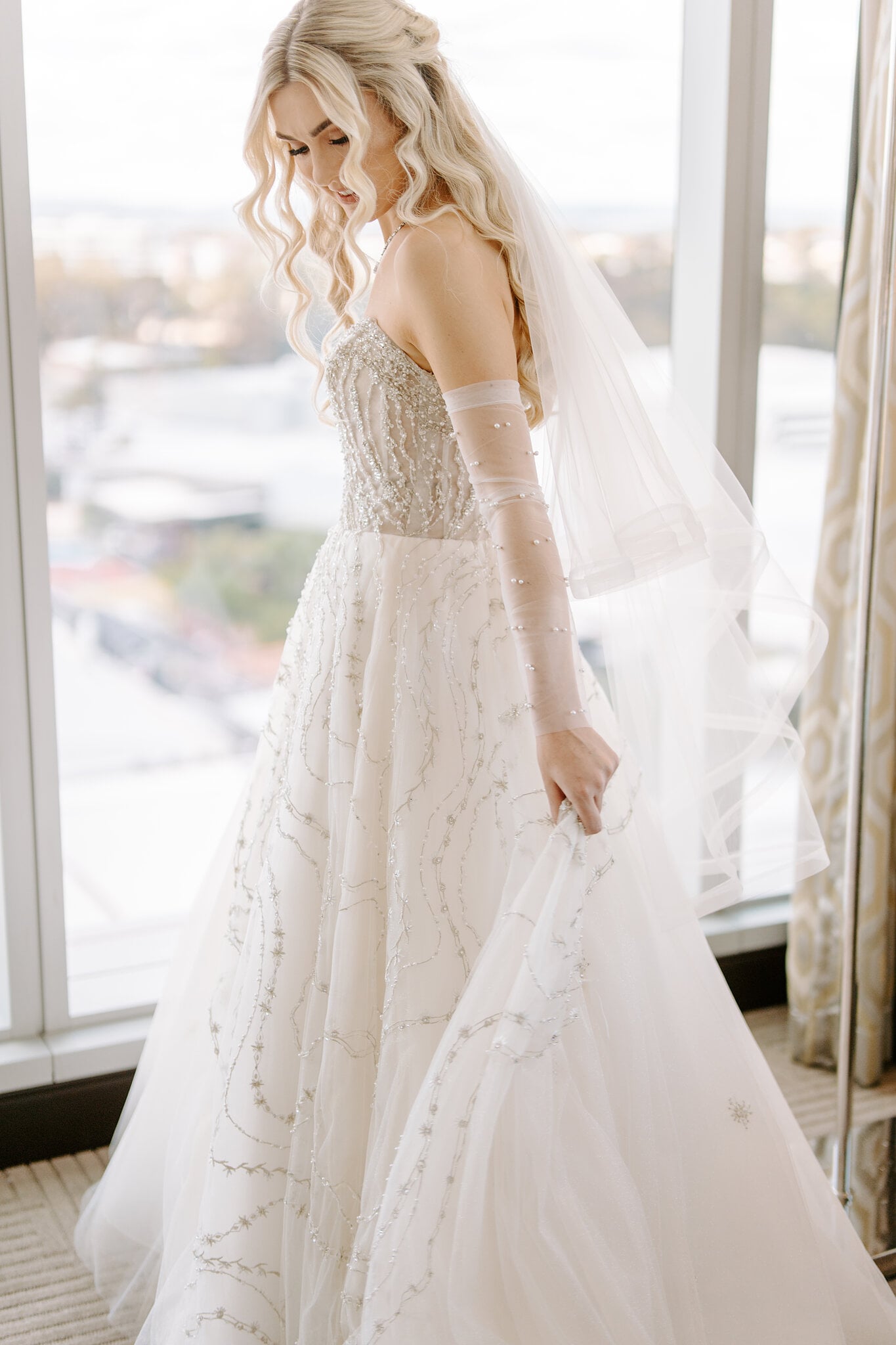 Claire lifts her beaded skirt by the window, sheer pearl sleeves on her arms.