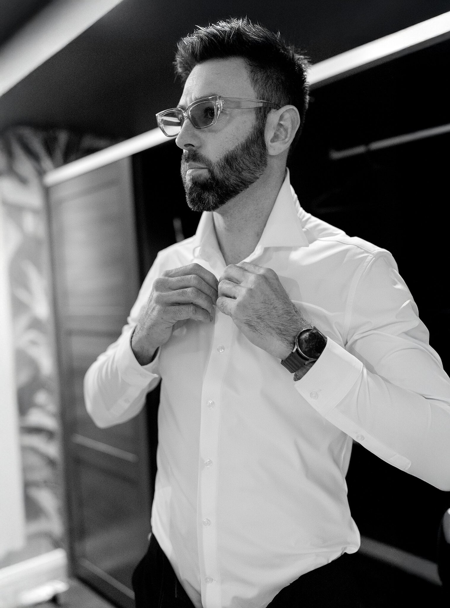 Dave buttons his white shirt by the window, glasses on, in black and white.