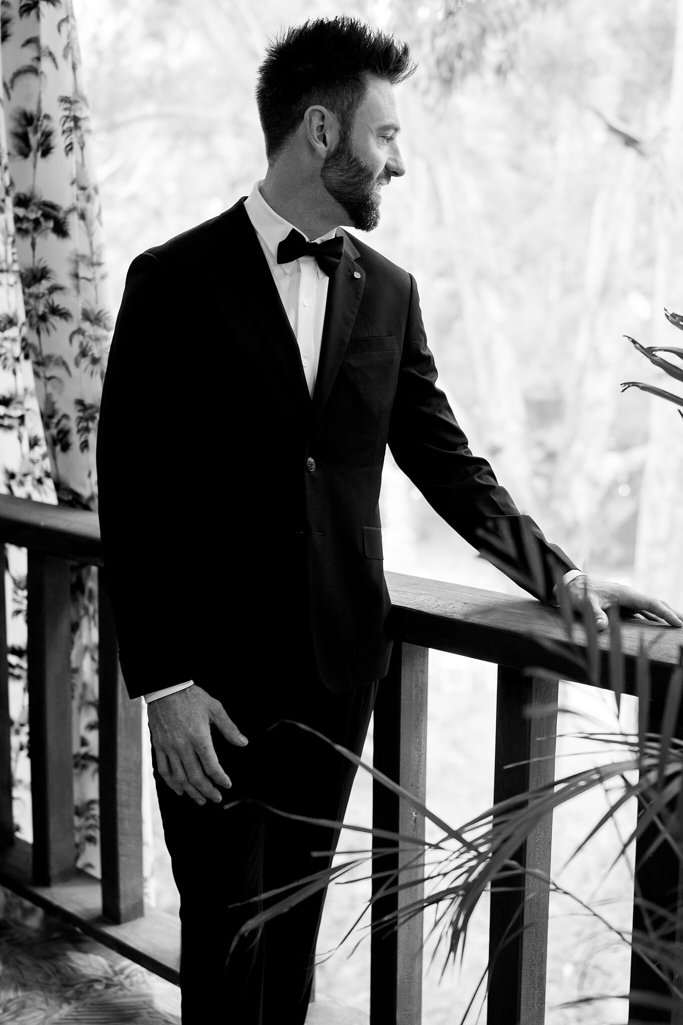 Dave leans on the balcony rail in his tuxedo, tropical leaves all around him.