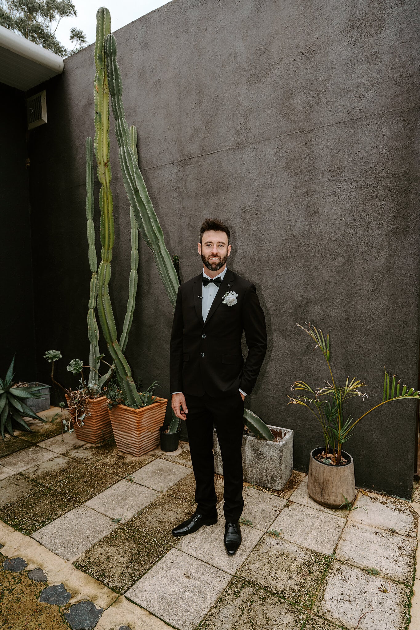 Dave stands beside a towering cactus against a charcoal wall in his tuxedo.