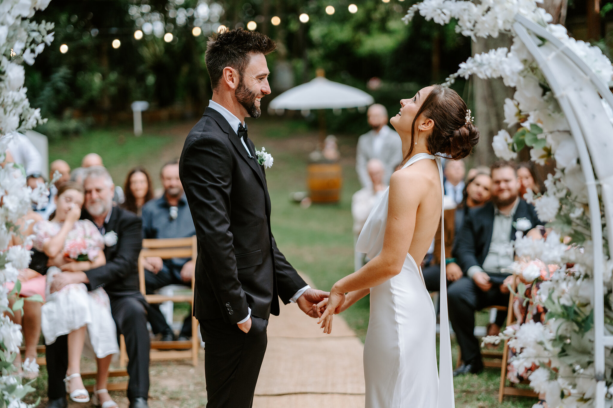 Emma laughs up at the sky as she and Dave hold hands under the white floral arch.