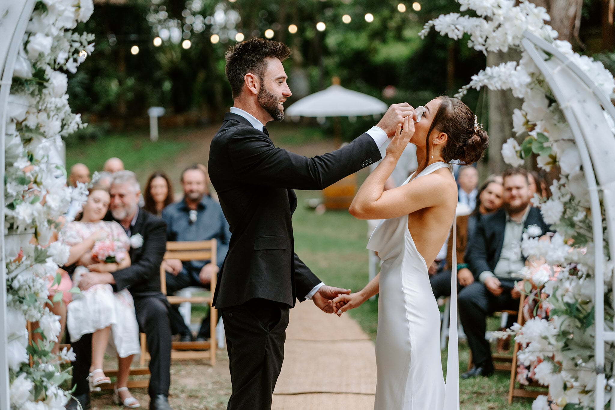Dave wipes a tear from Emma’s cheek during the vows as guests watch from timber chairs.