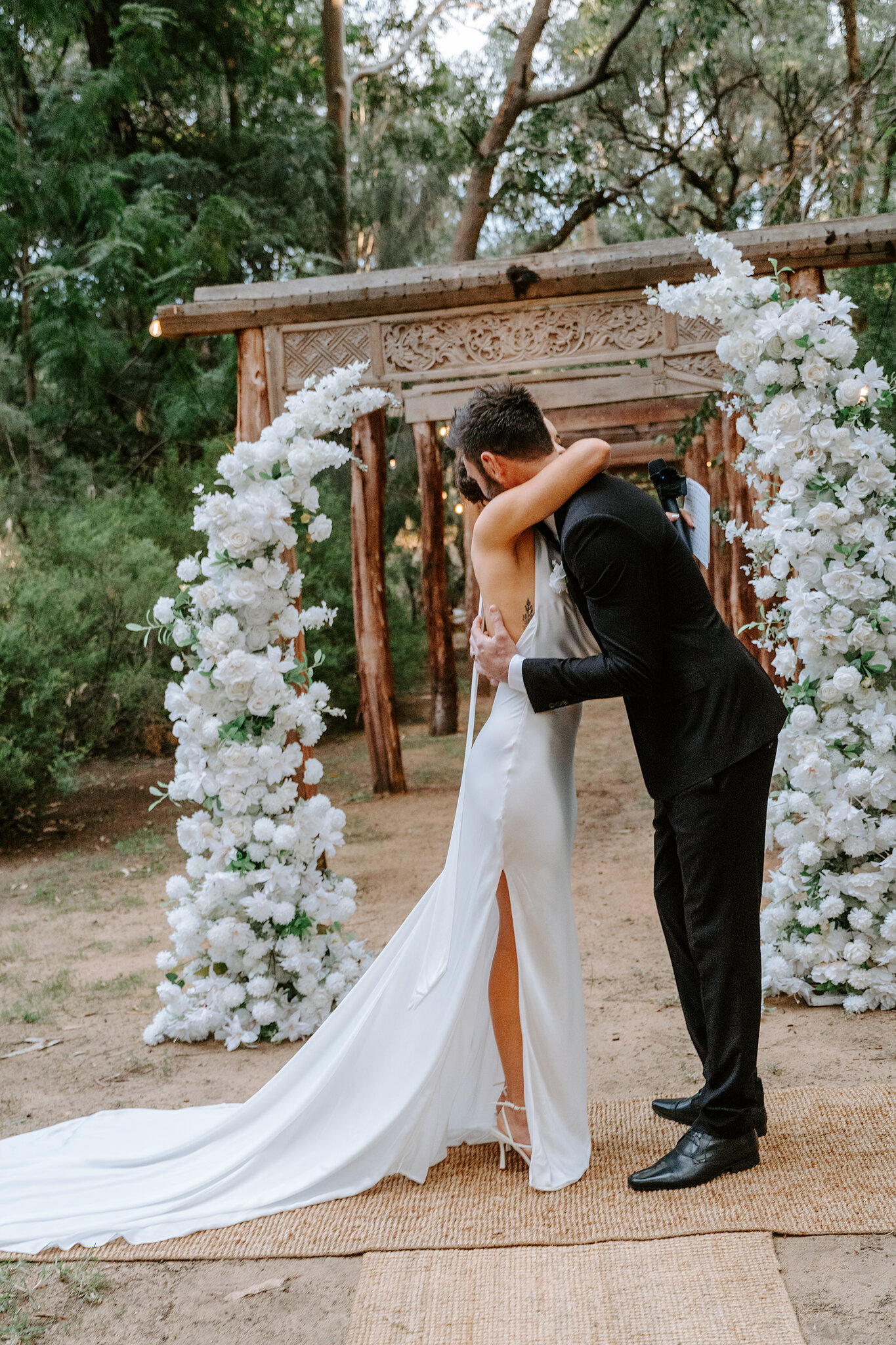 Emma and Dave kiss under the white floral arch and carved timber pergola.