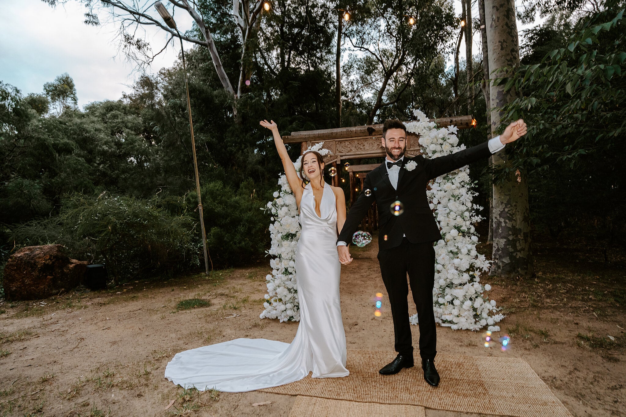 Emma and Dave throw their arms in the air under the floral arch, just married.