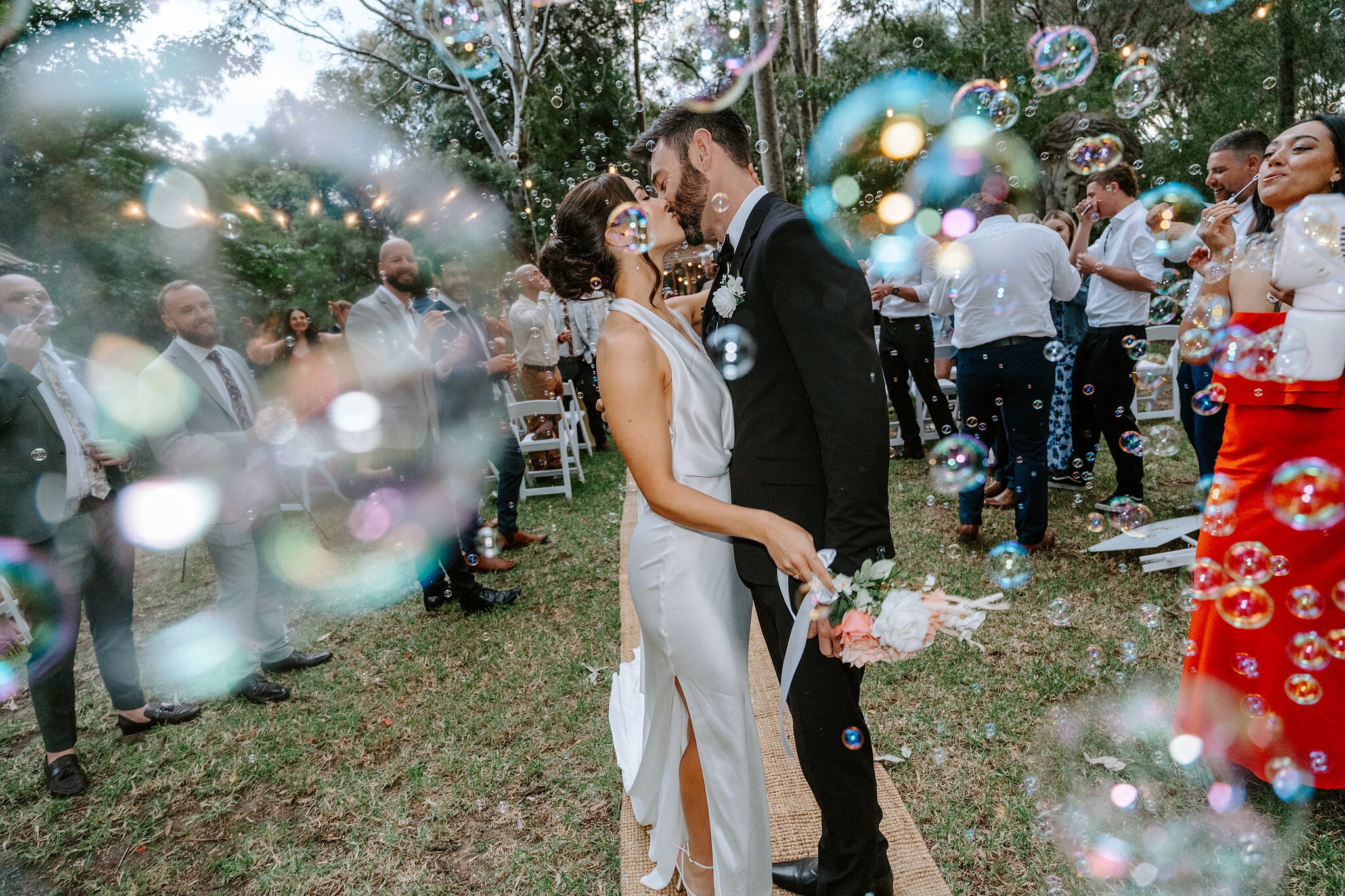 Soap bubbles blur the foreground as Emma and Dave kiss among their guests.