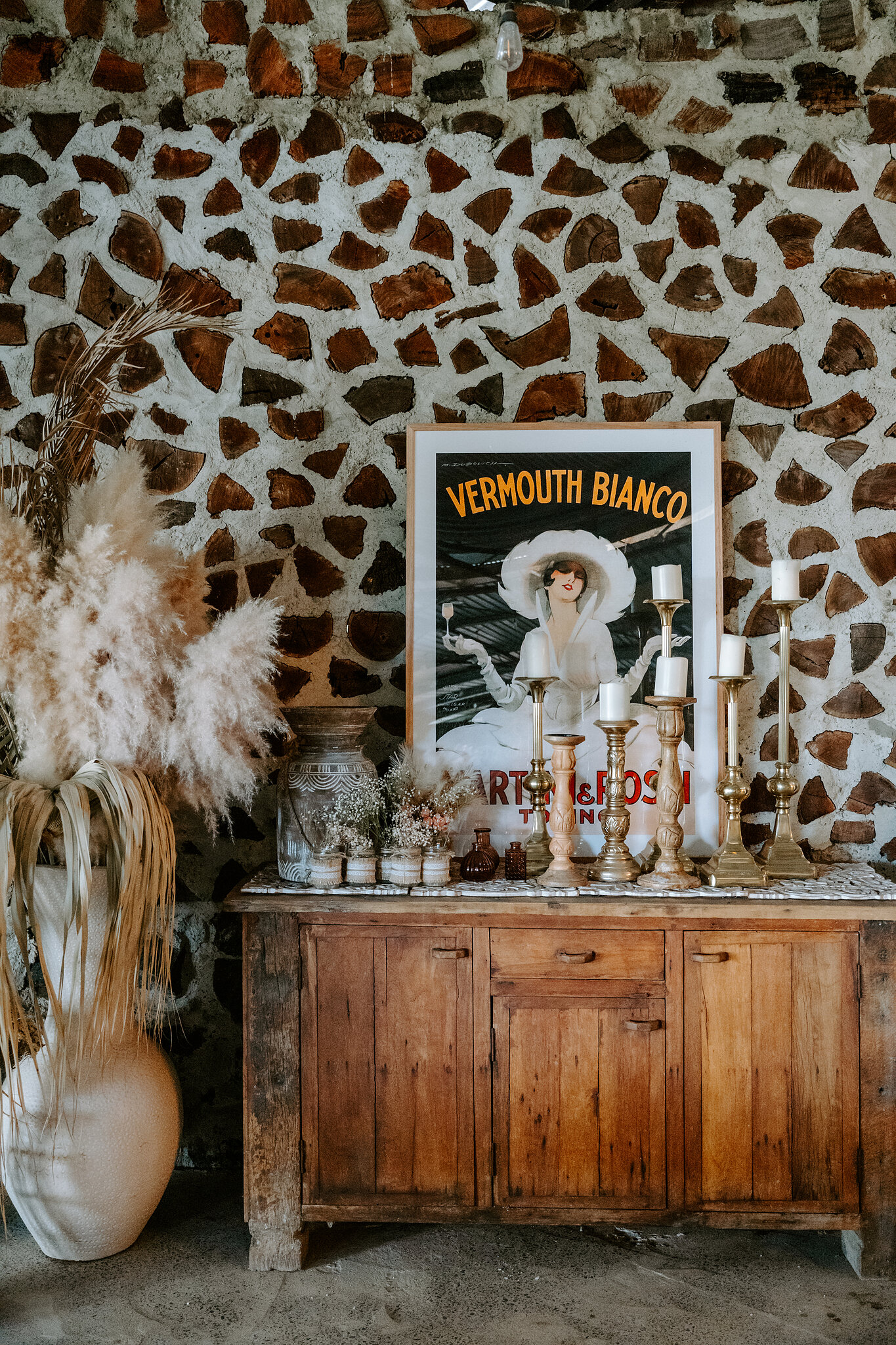 Pampas, candlesticks and a vintage Vermouth Bianco poster on a timber dresser against a stone wall.
