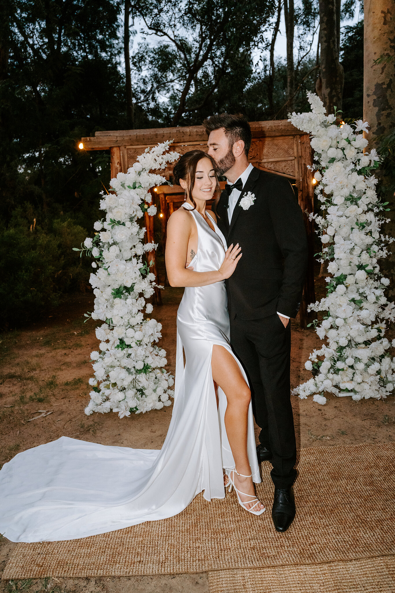 Emma and Dave hold each other in front of the white floral arch at dusk.