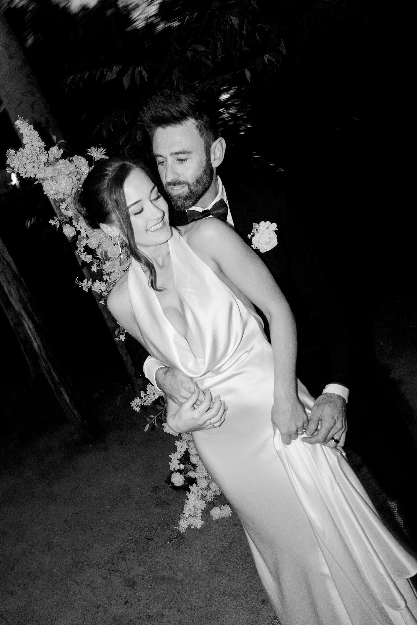 Emma laughs in Dave’s arms beside the floral arch at night, in black and white.