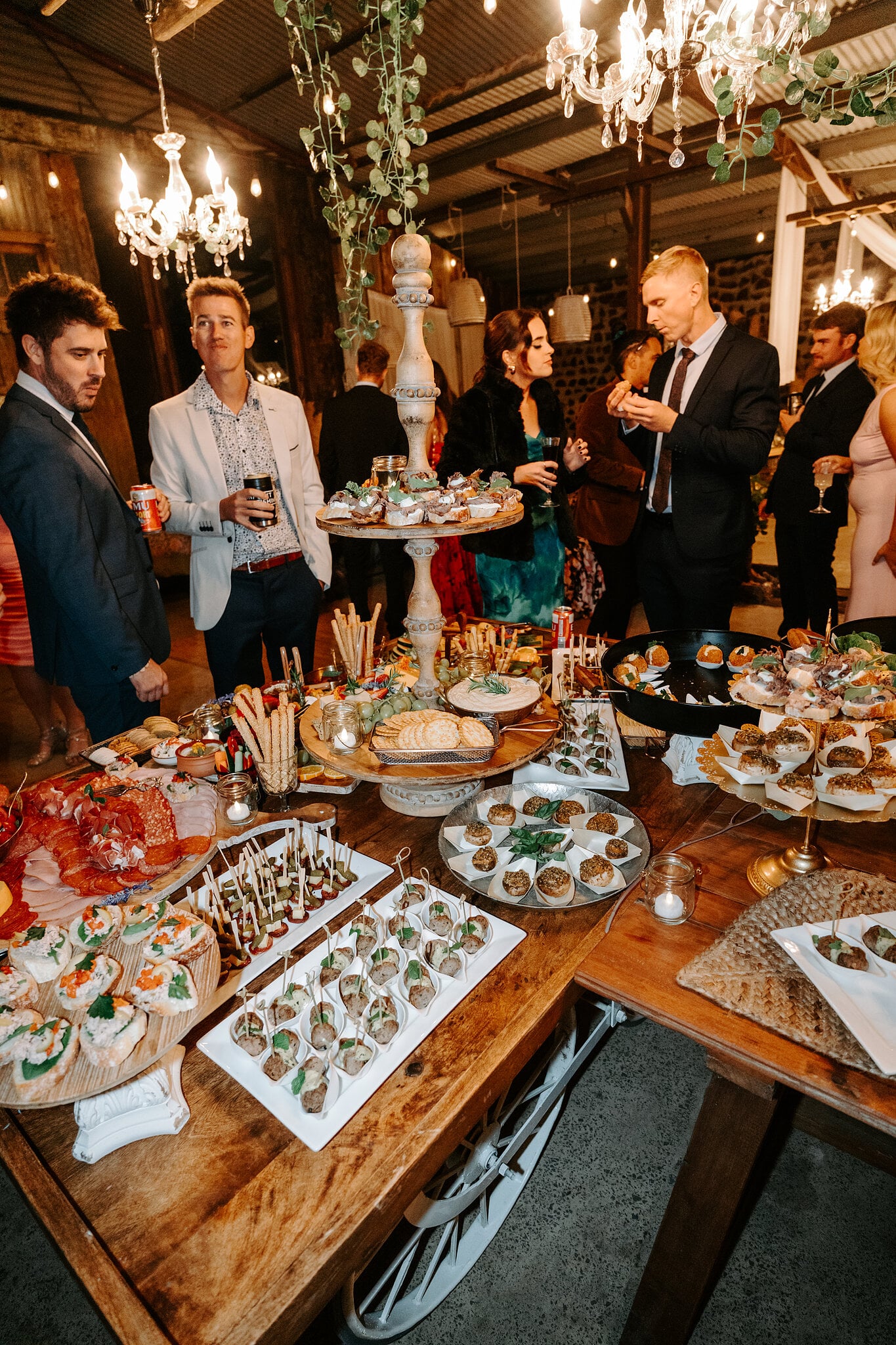 Guests gather round a long grazing table under chandeliers in the rustic shed.