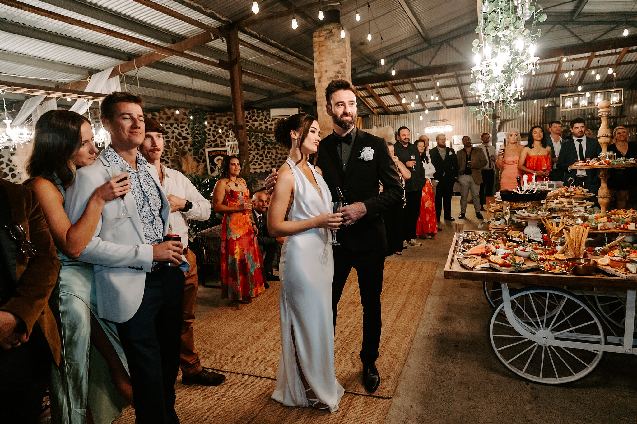 Emma and Dave mingle with guests in the shed beside a grazing cart and chandeliers.